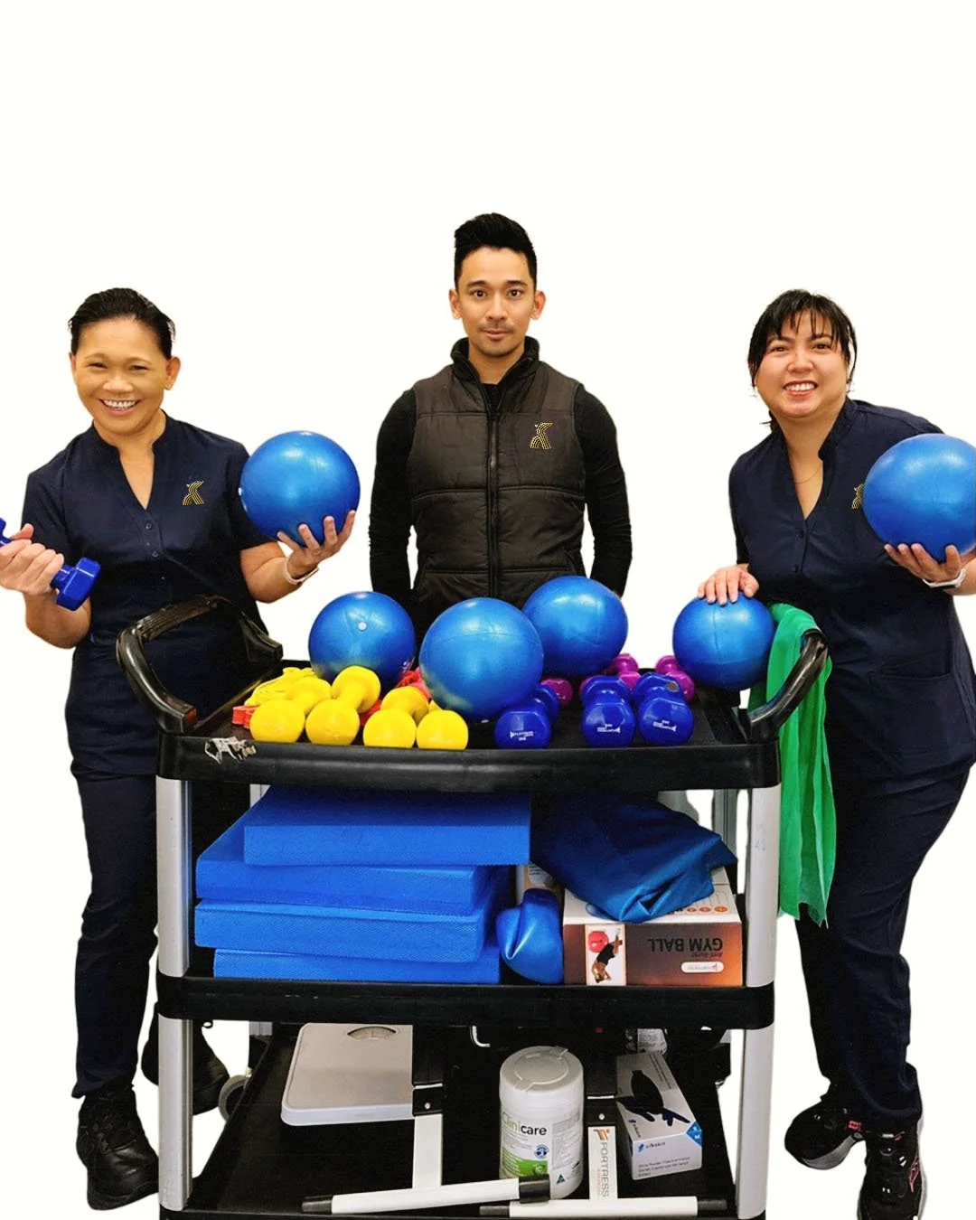 Three people standing behind a fitness equipment cart loaded with exercise gear, including blue exercise balls, small yellow dumbbells, and blue dumbbells, in a fitness center.