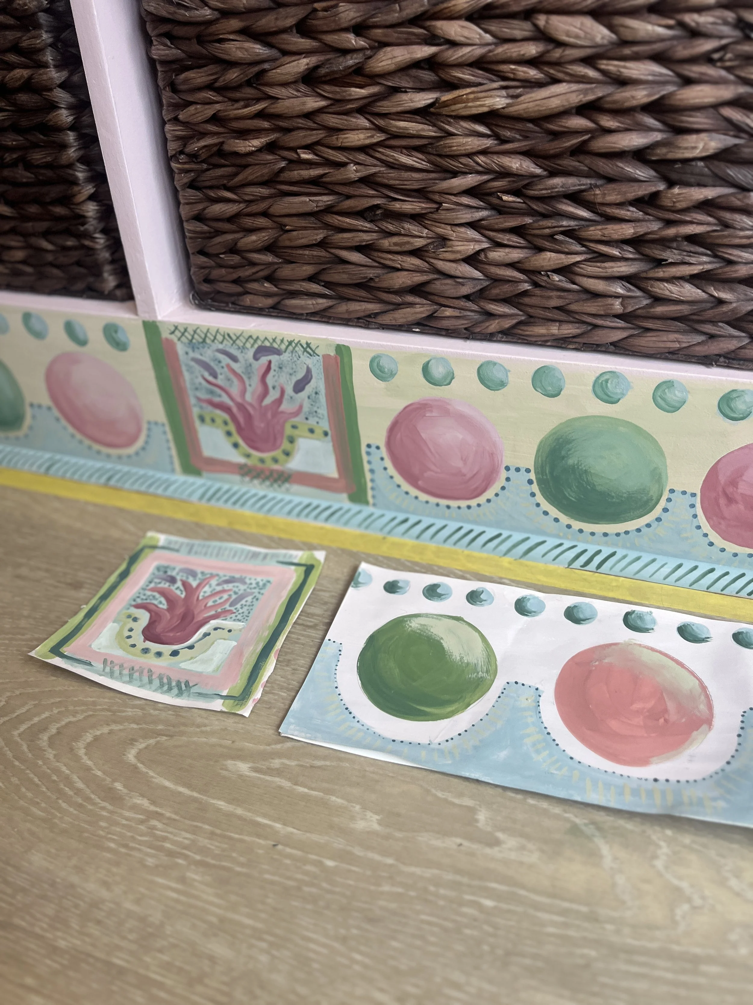 Hand-painted artwork featuring a pink plant or abstract shape with dotted border, placed on a table alongside matching painted sheets of paper, with a decorative painted border on the wall in the background.