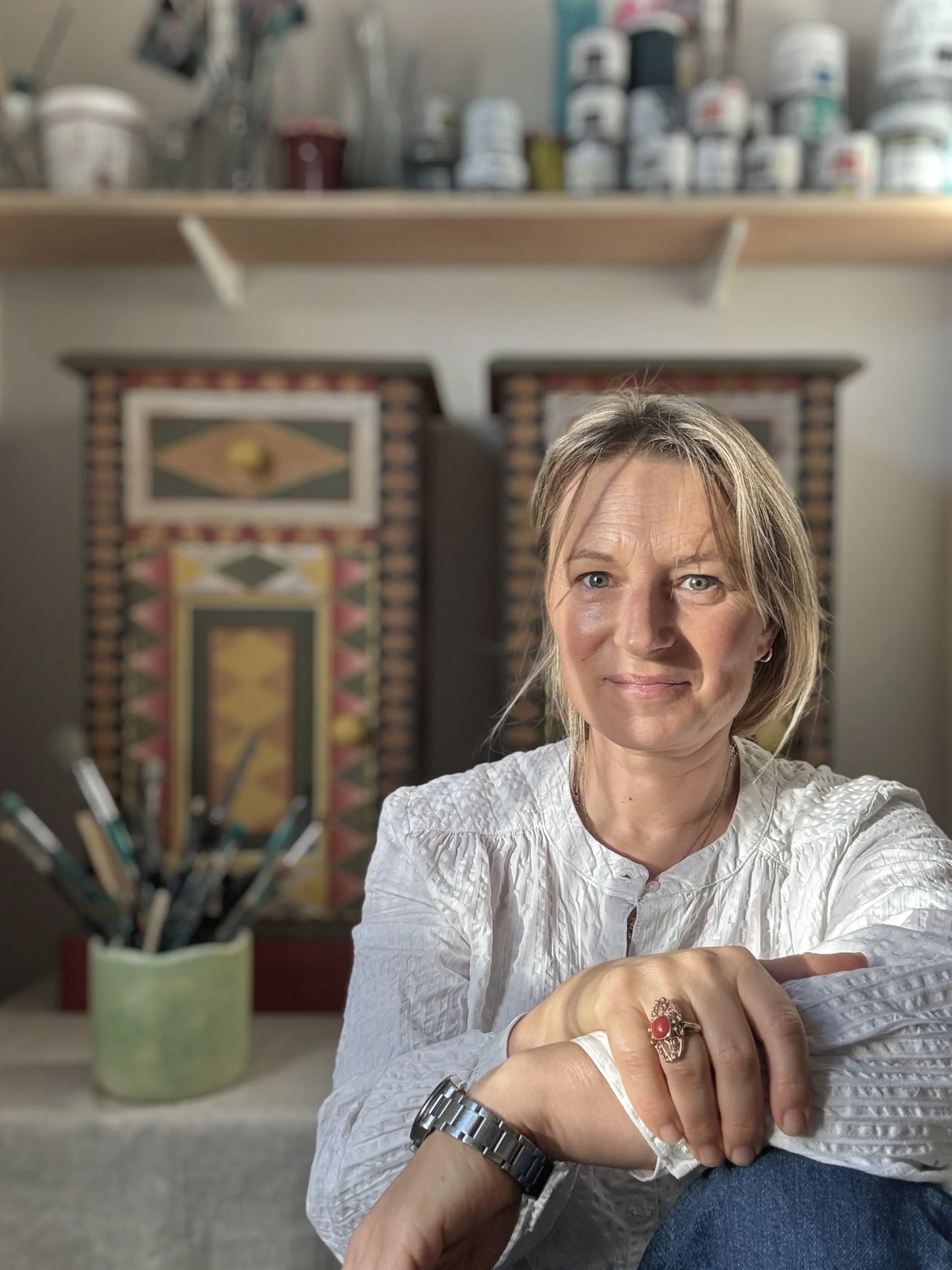 A woman with blonde hair, wearing a white blouse and a wristwatch, sitting indoors in front of a colorful wooden cabinet with artwork and a container of paintbrushes, looking at the camera.