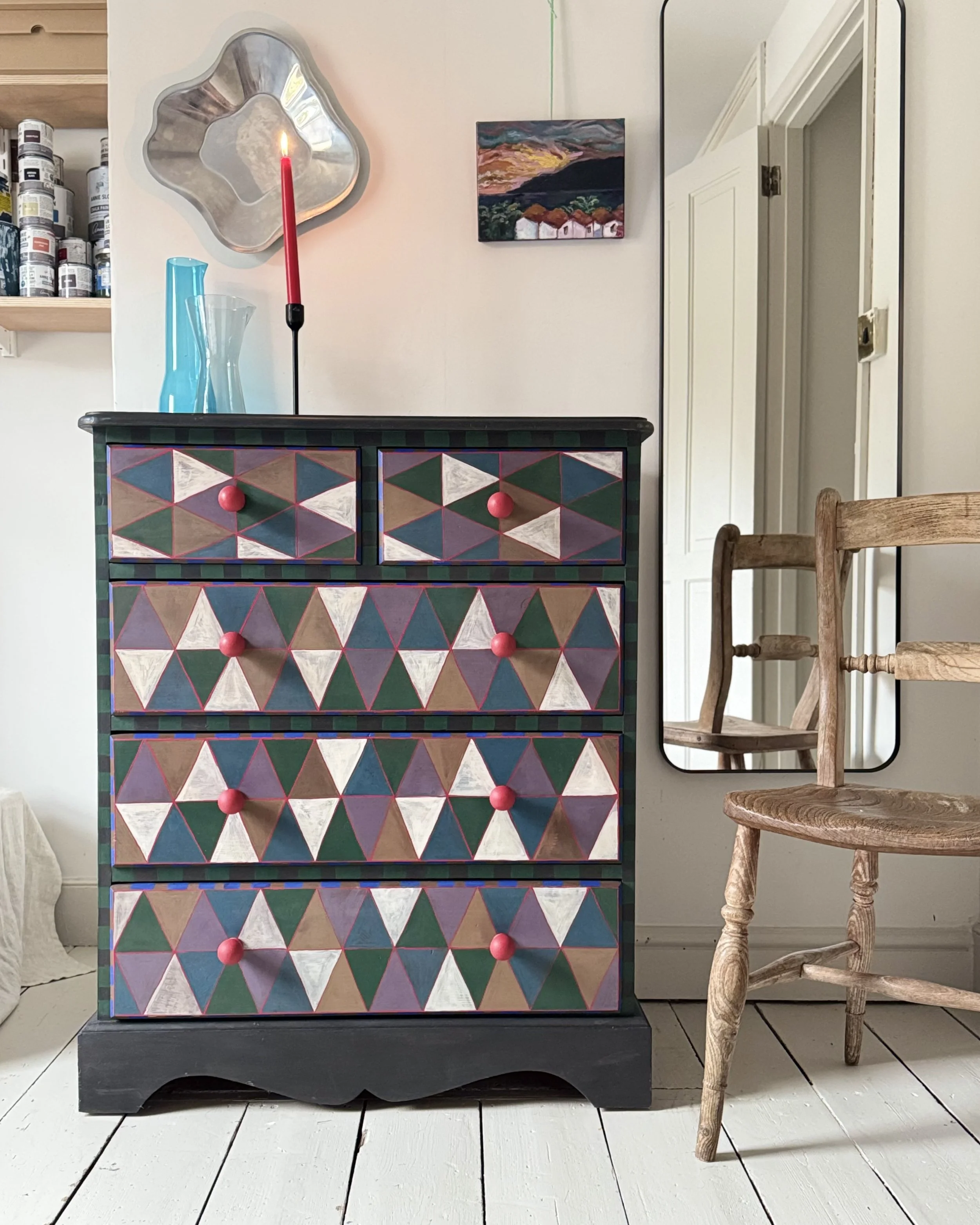 Colorful dresser with geometric triangle pattern and pink knobs, a tall candle in a holder, and a mirror on the wall in a cozy room.
