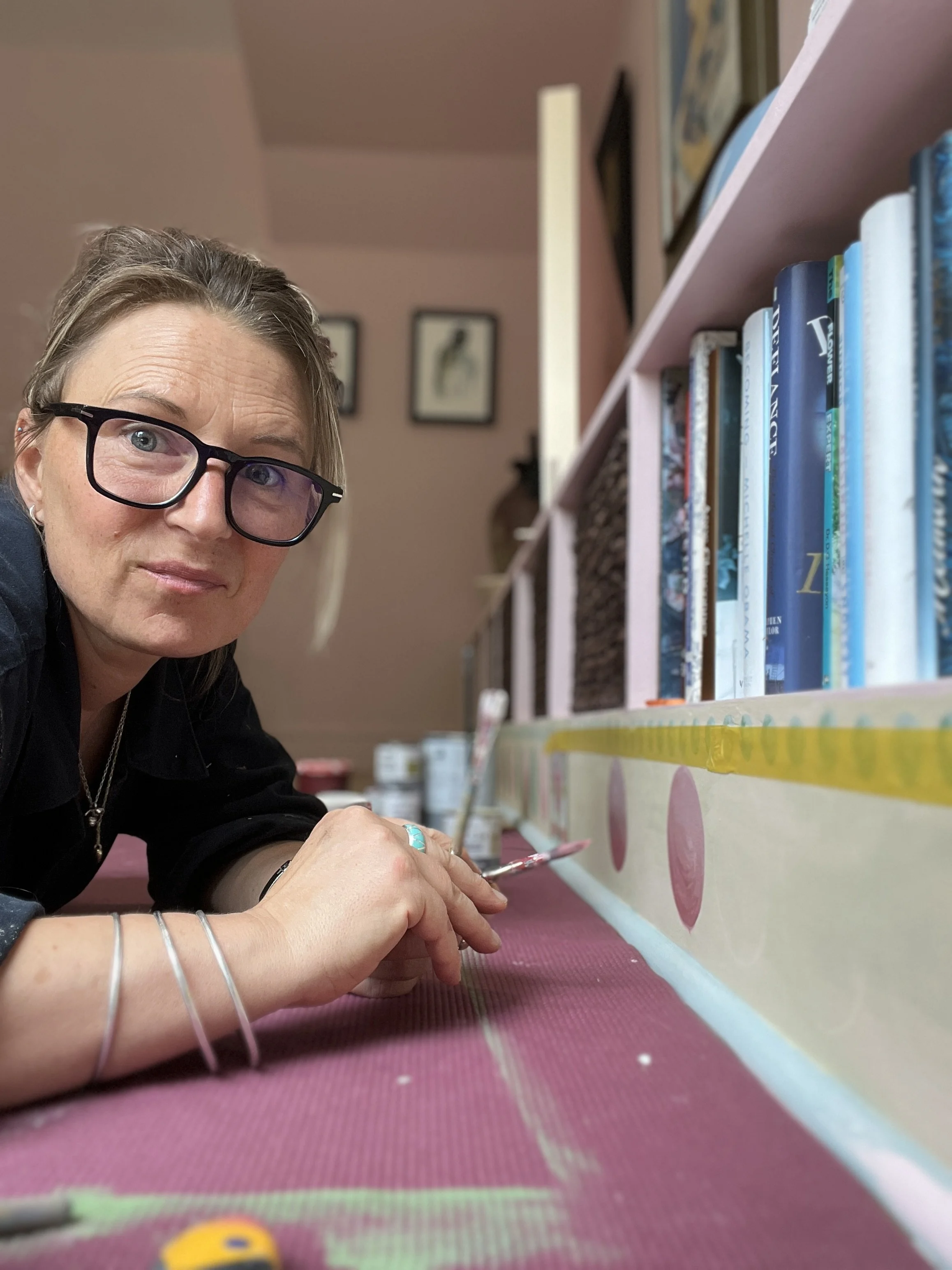 A woman with glasses, jewelry, and a black shirt leans over a pink table, painting a decorative border along a bookshelf filled with colorful books in a cozy room.