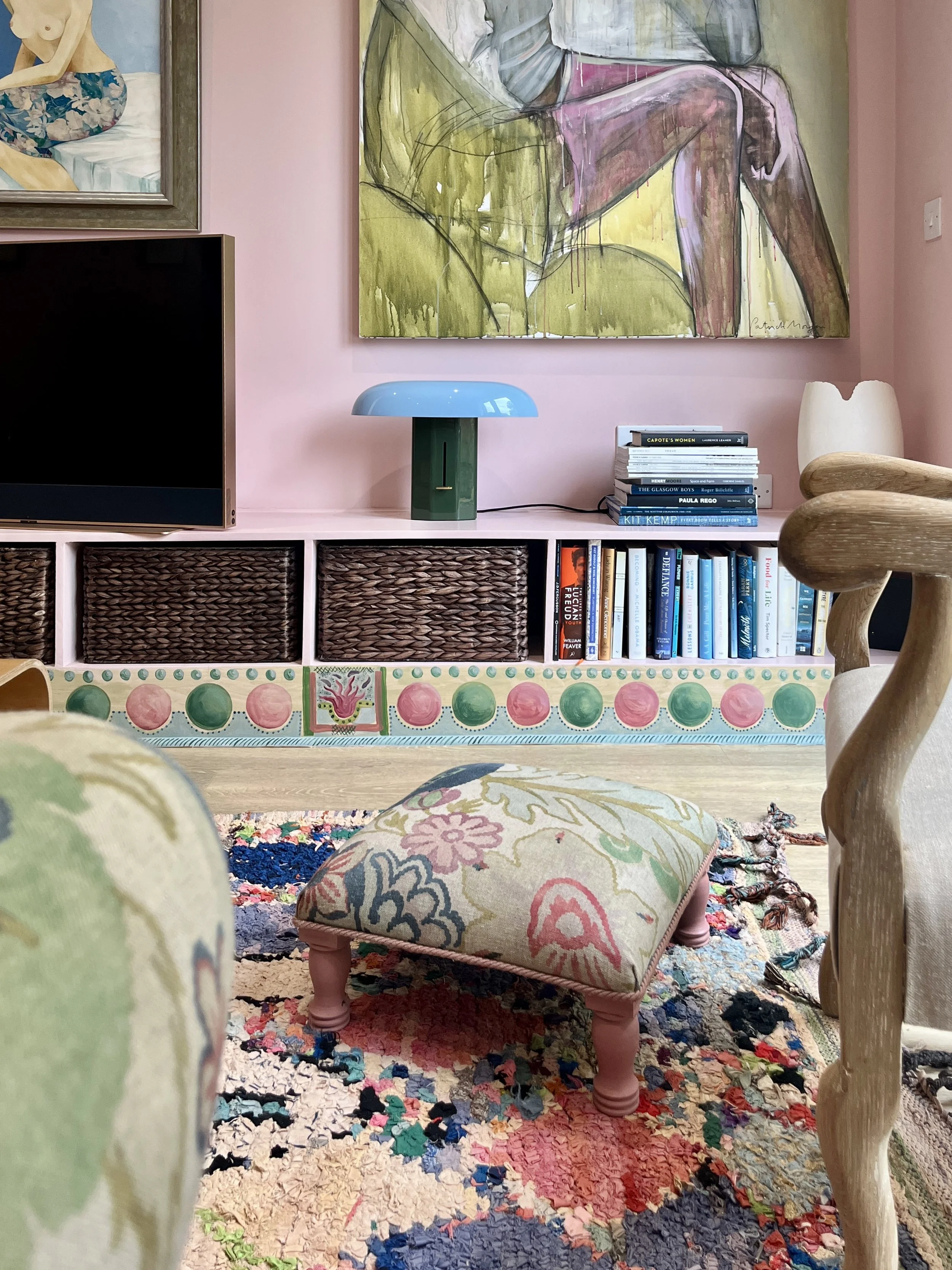 Interior living room with pink walls, a pink and multicolored artistic rug, a small cushioned footstool with floral fabric, a pink and green shelf with books, a blue lamp with a green base, a flat-screen TV, and colorful paintings.