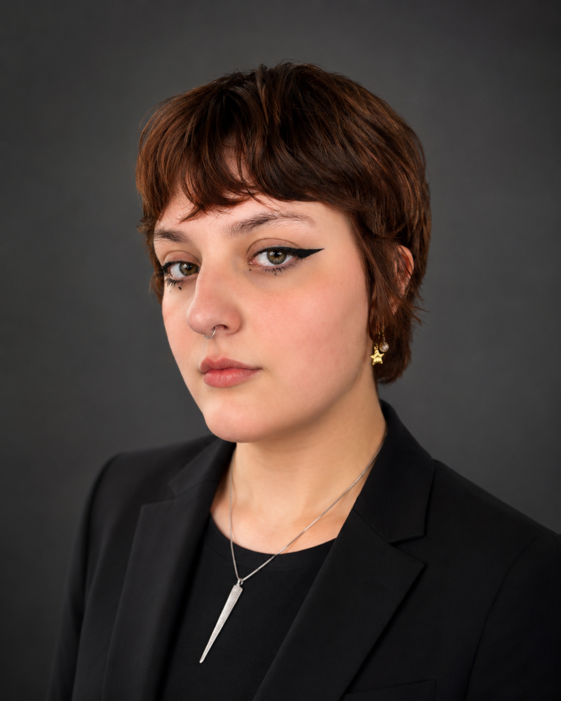 A young woman with short, reddish-brown hair and light skin, wearing makeup with winged eyeliner, a septum piercing, a star-shaped earring, and a silver pendant necklace, dressed in a black blazer against a dark gray background.