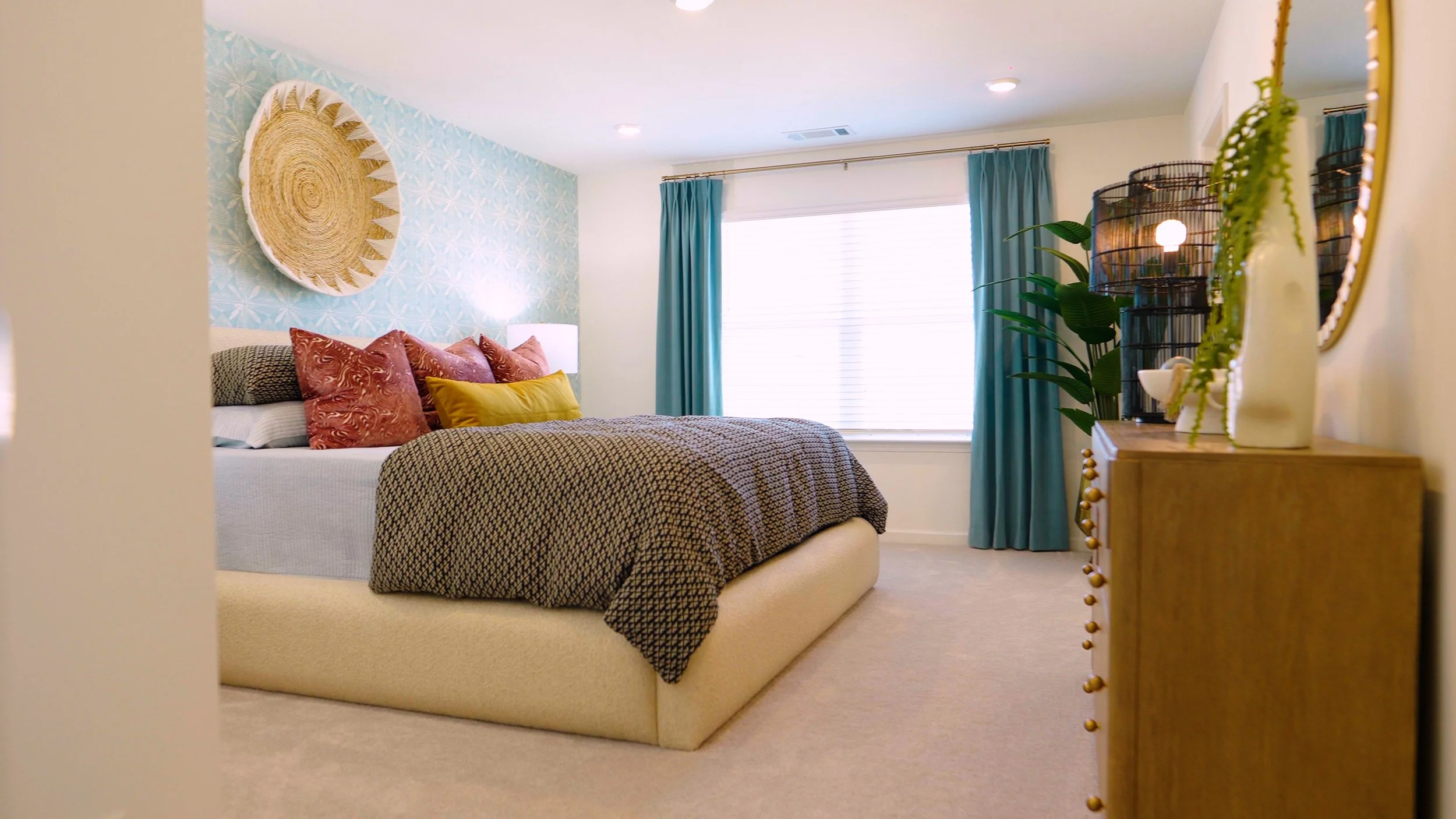 A bedroom with a bed, colorful pillows, a dresser with decorative items, a large window with blue curtains, and wall art.