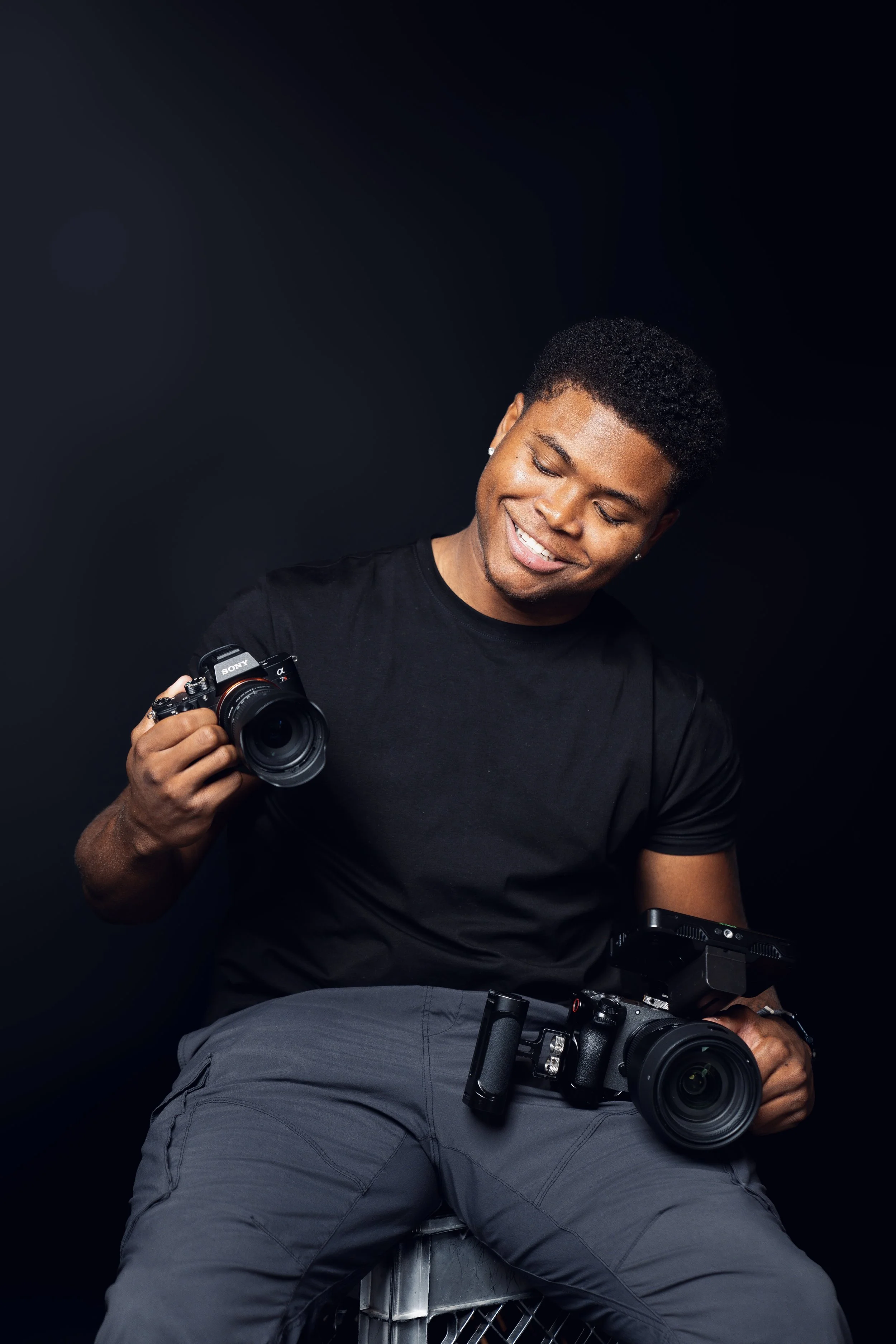 A young man in a black t-shirt and gray pants holding two cameras, smiling and looking down against a dark background.