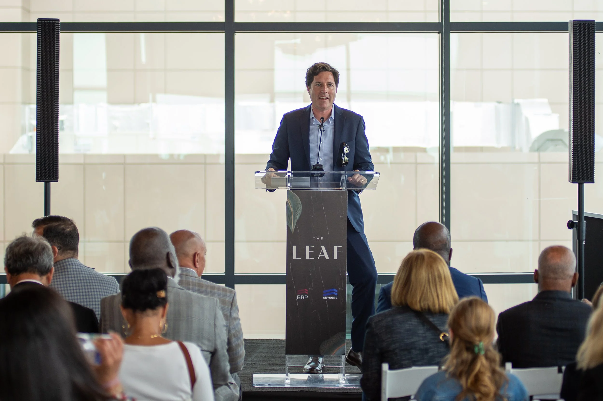 A man speaking at a podium with a sign that reads 'The LEAF' in a room with large windows, surrounded by an audience.