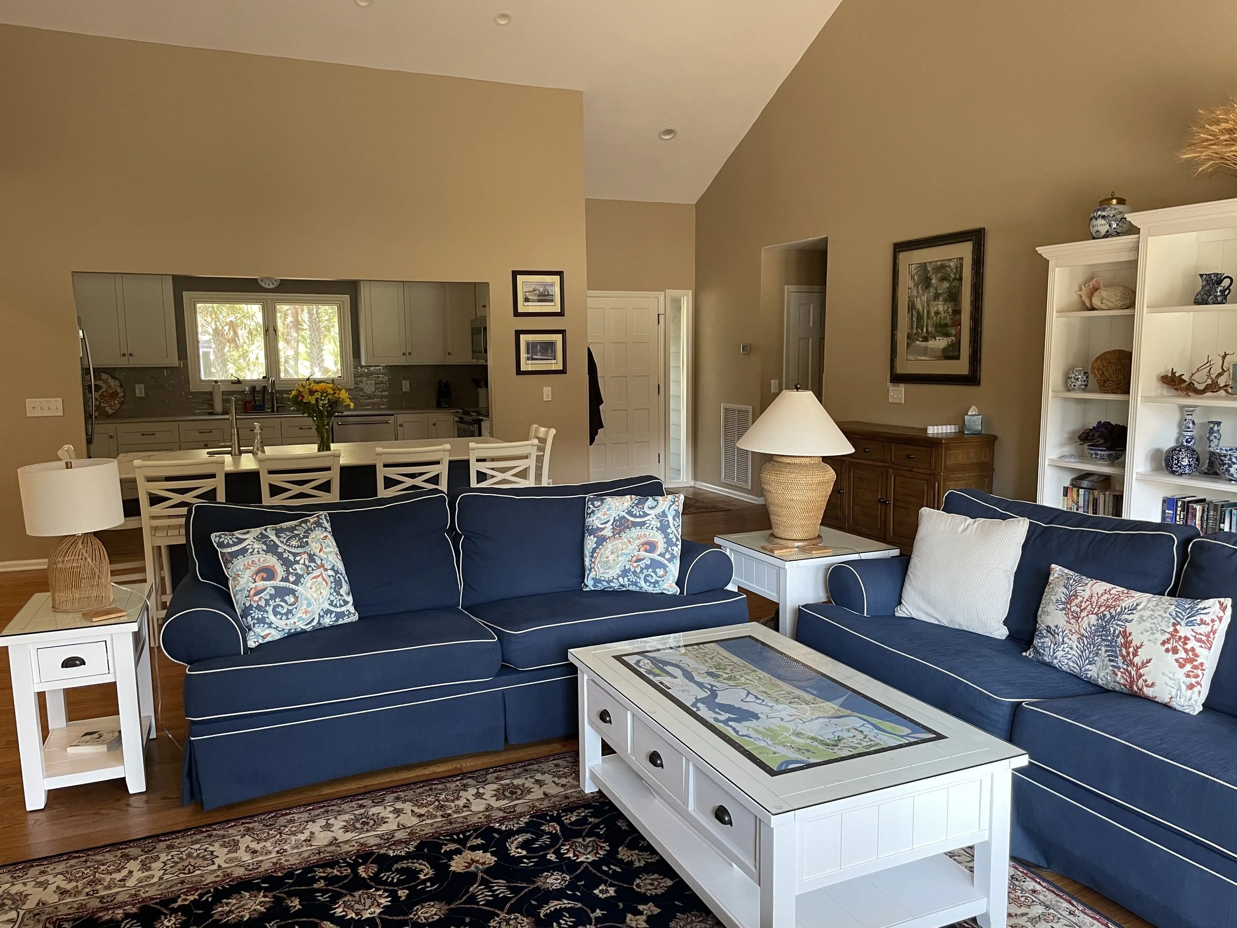 Living room with blue sofas, white side tables and a white coffee table with a map, bookshelf with blue and white vases, kitchen with white cabinets, dining area with white chairs, and beige walls.