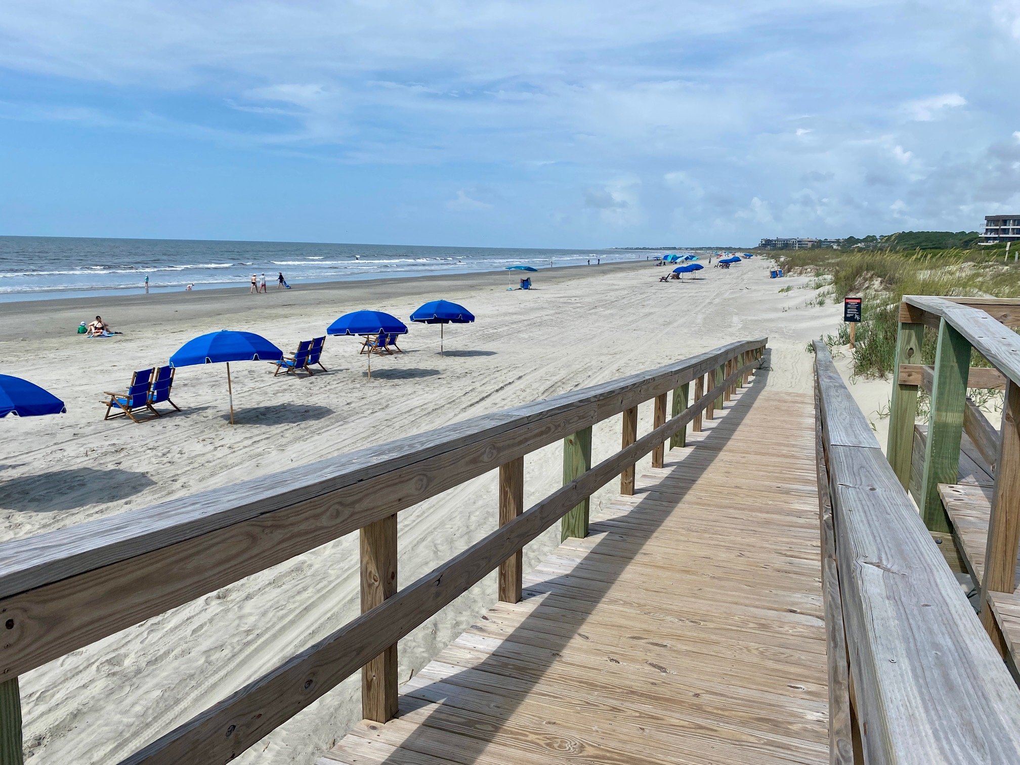 Wooden boardwalk leading to a sandy beach with blue umbrellas and beach chairs, people walking, and ocean waves under a partly cloudy sky.