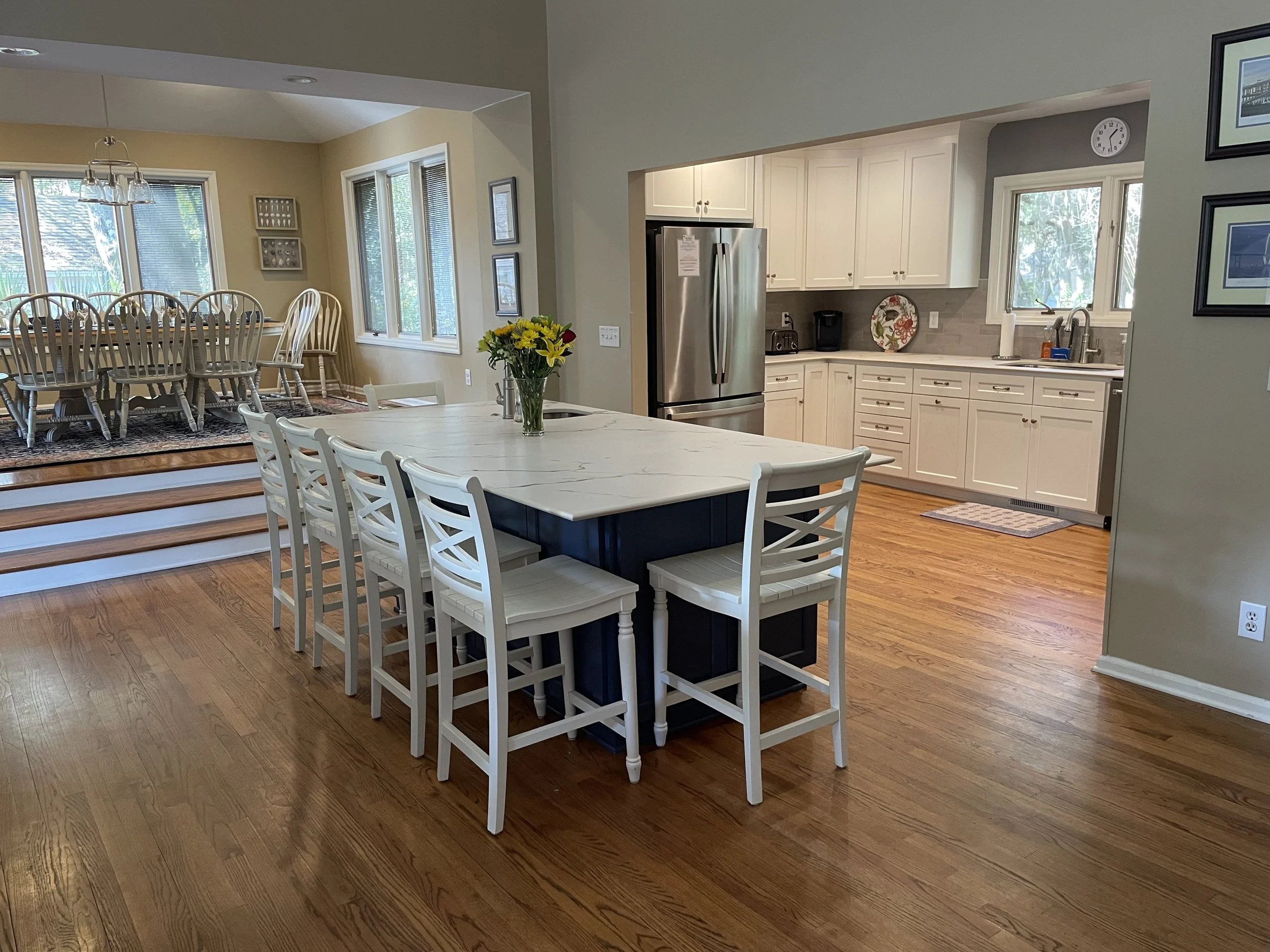 Open-concept kitchen and dining area with hardwood floors, a white kitchen with multiple cabinets, a stainless steel refrigerator, a small kitchen island with white chairs, and a formal dining table with matching chairs on a raised platform with step