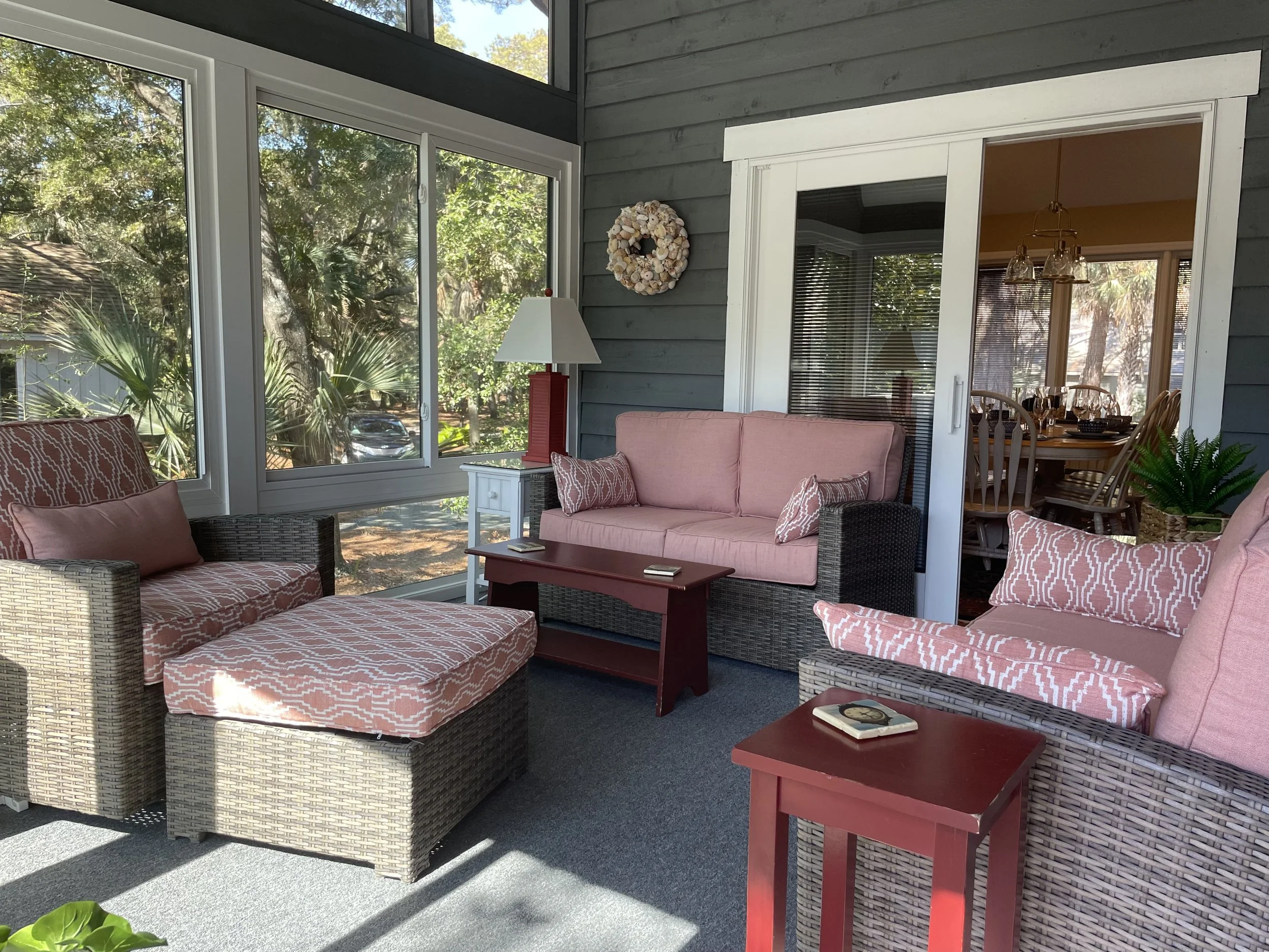 Sunroom with wicker furniture, pink cushions, and red side tables, adjacent to a dining area with wooden chairs, decorated with plants and natural light.
