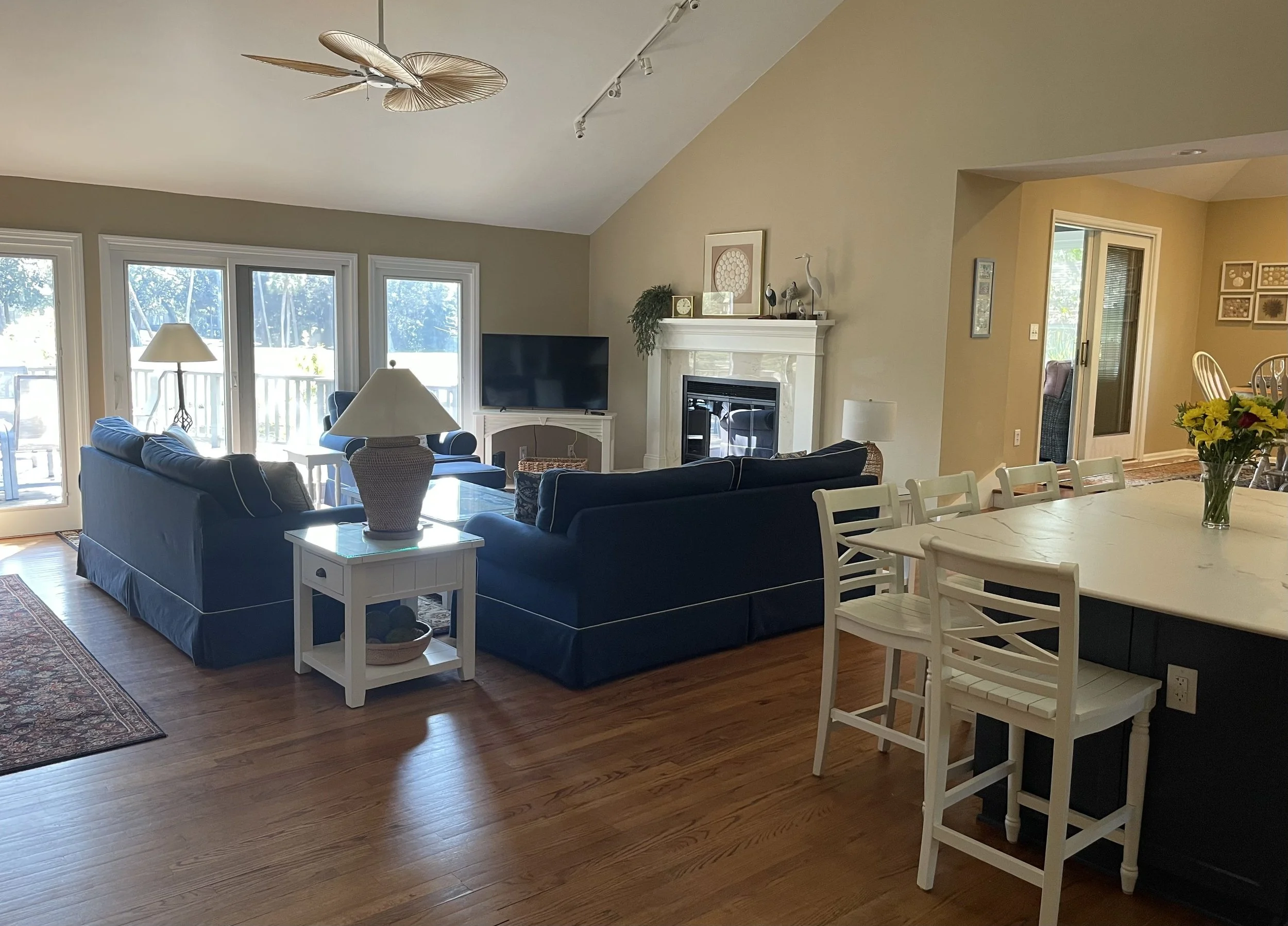 Living room with navy blue sofas, white side table with lamp, fireplace, flat-screen TV, hardwood floors, and a patio door leading outside. To the right is a dining area with a white table, chairs, and a vase of yellow flowers.