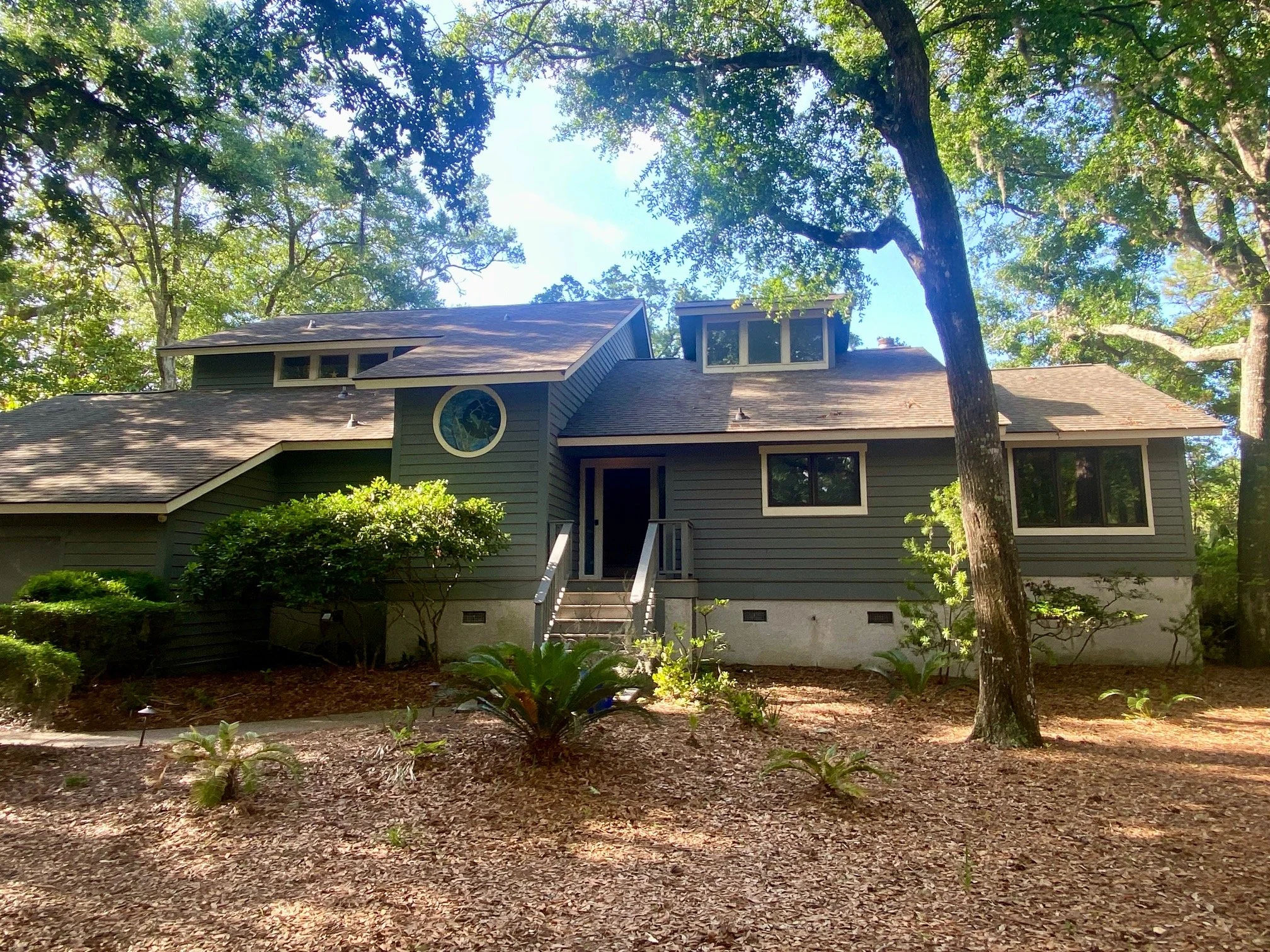 A two-story house painted in green, surrounded by trees and shrubs, with a staircase leading to the front door.