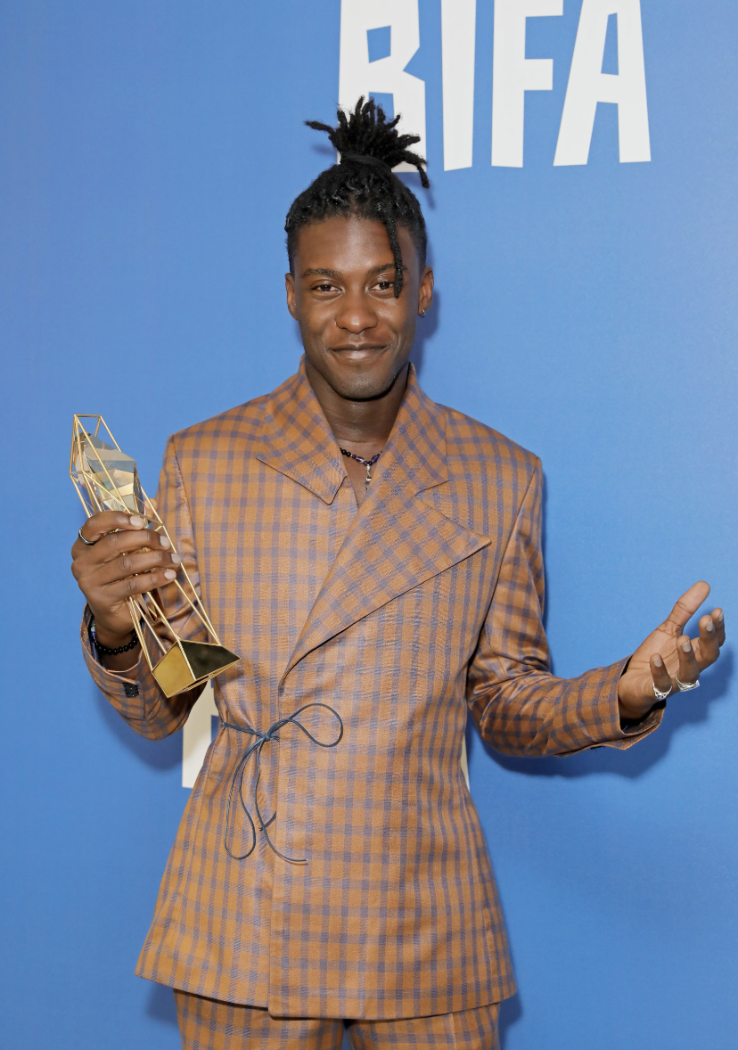 A man with dreadlocks in a brown checkered suit holding a trophy at an awards event with a blue background and the letters 'BIFA' visible behind him.