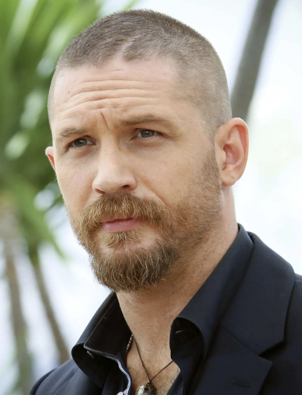 Close-up of a man with short hair and a beard wearing a black shirt and necklace, with a blurred outdoor background.