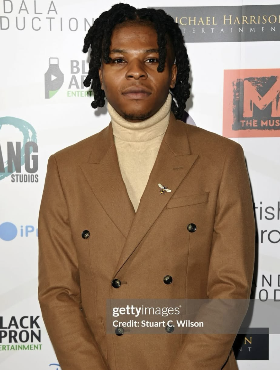 A man with dark skin and black dreadlocks wearing a tan double-breasted blazer with a bee pin on the lapel and a beige turtleneck shirt, standing in front of a step and repeat background with logos.