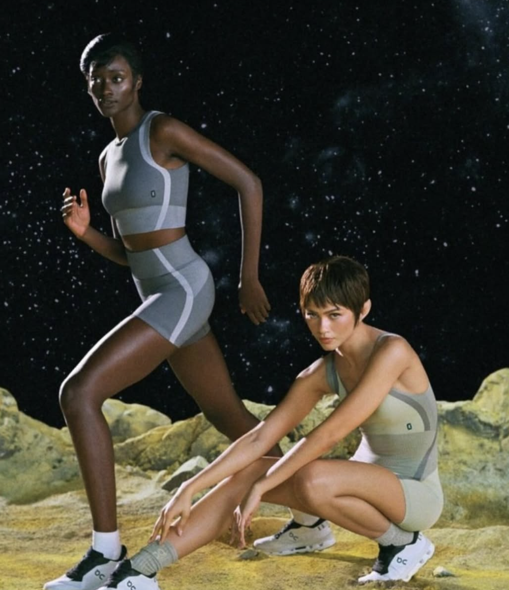 Two women in athletic outfits on a rocky terrain with a starry space background, one standing and one crouching, dressed in sporty, futuristic attire.