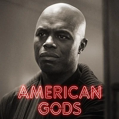 A black and white close-up image of a serious man with a shaved head, wearing dark clothing. The text "American Gods" in red neon font is at the bottom of the image.