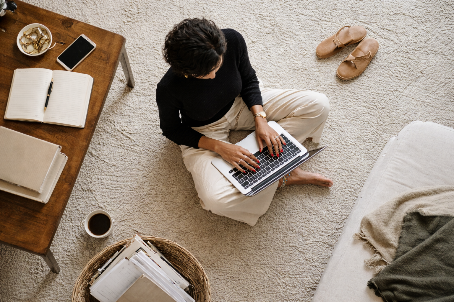 A woman sitting cross-legged on a carpeted floor working on a laptop, with an open notebook, a pen, a smartphone, a cup of coffee, and a basket of papers nearby, and a pair of shoes on the floor.