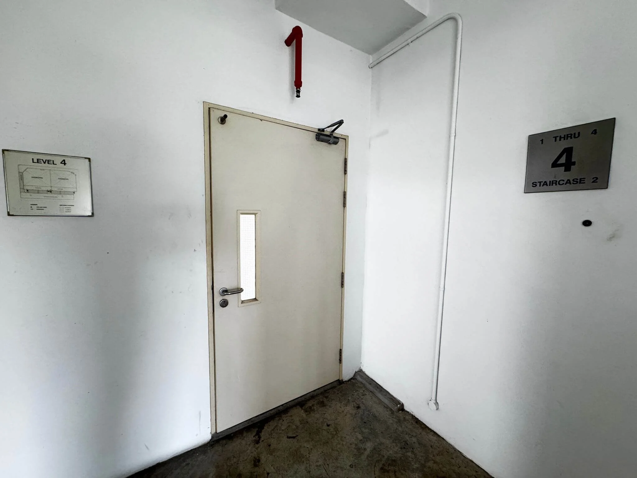 White stairwell door with small window, surrounded by white walls with signs indicating Level 4 and Staircase 2, and a red fire alarm with the number 1 above.