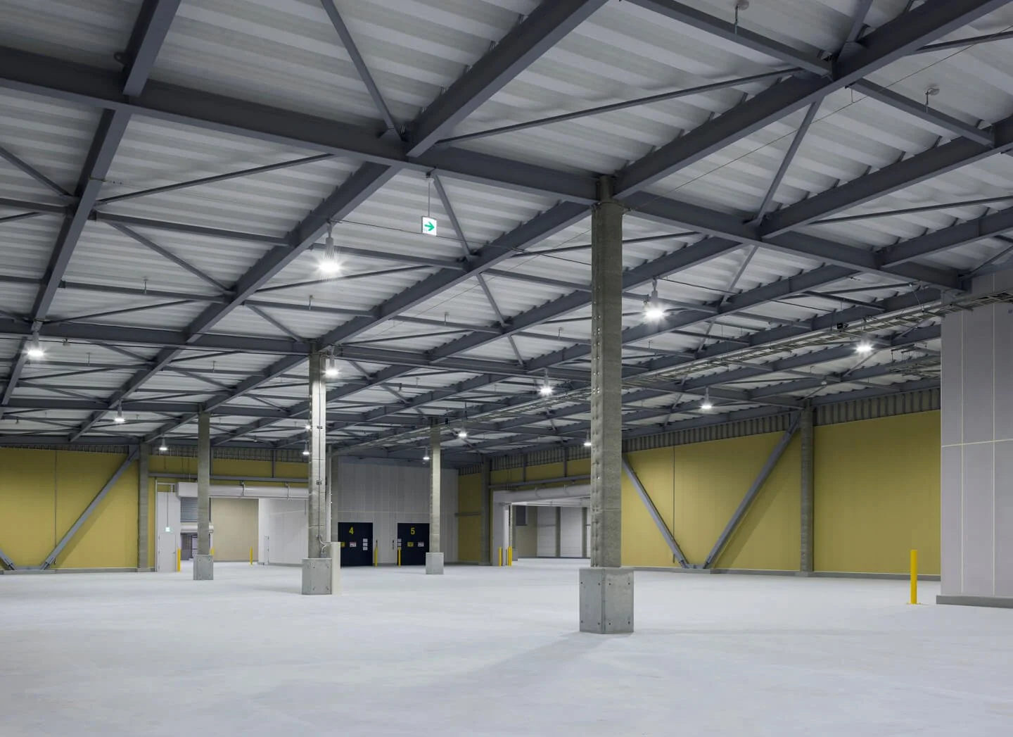 Empty indoor warehouse space with high ceiling, metal roof, concrete support columns, yellow wall sections, and overhead lighting.