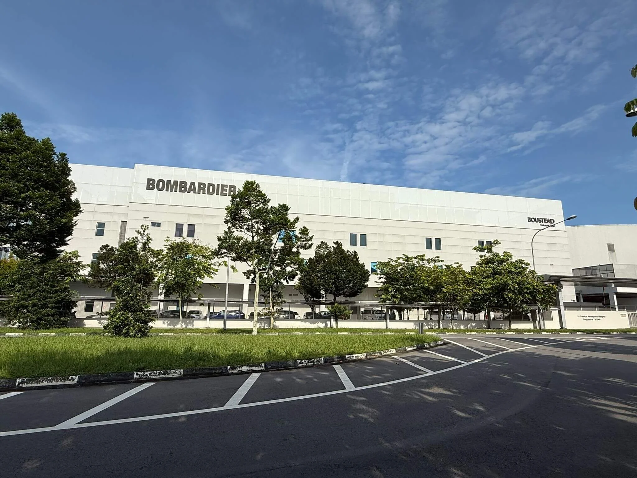 Exterior of a large white building with the name 'Bombardier' on the front, surrounded by trees and a parking lot under a partly cloudy sky.