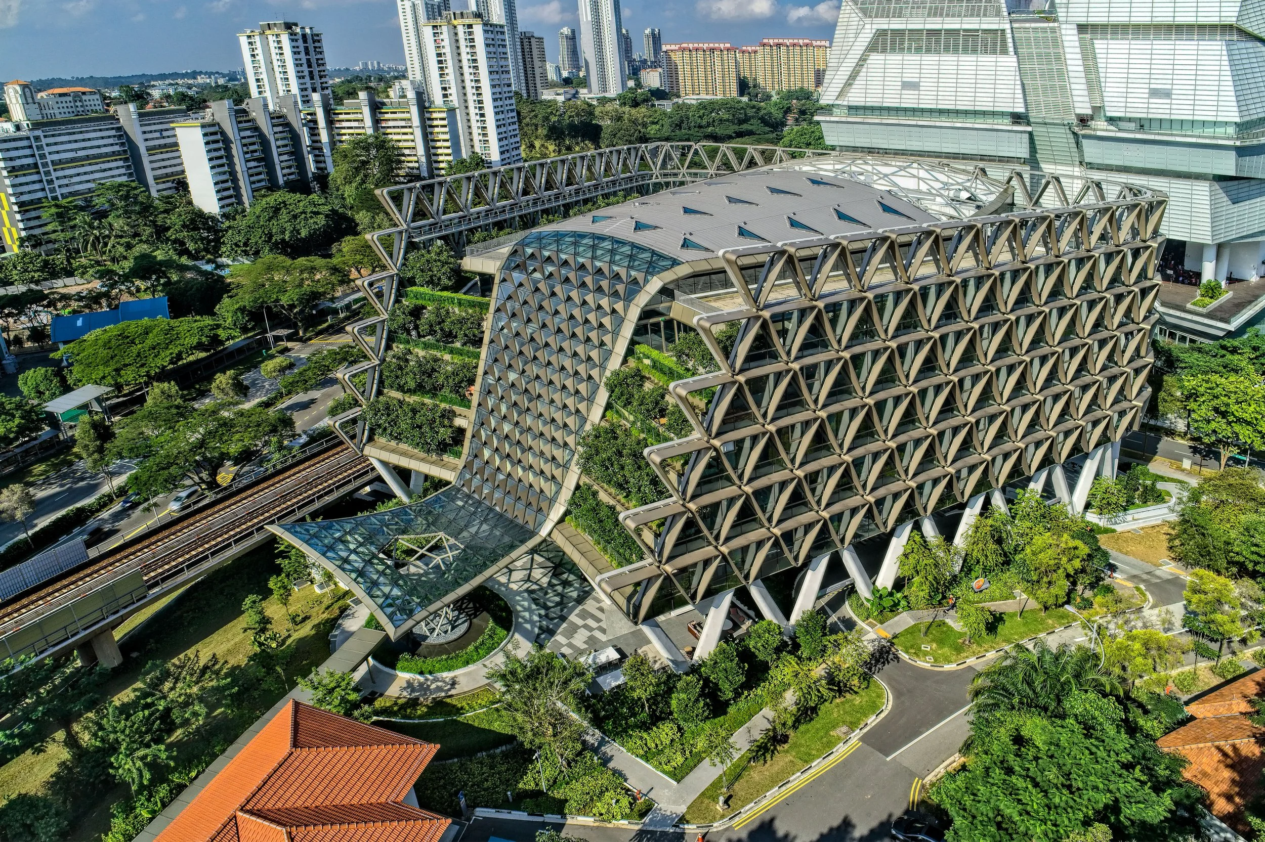 A modern architectural building with a unique curved design and a lattice-like exterior, surrounded by lush green trees and landscaped areas in an urban cityscape.
