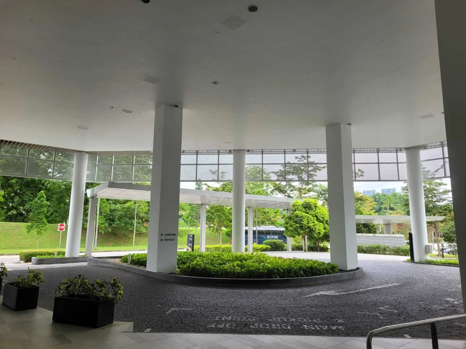 Covered hotel or office building entrance with white columns, green landscaping, and glass windows, with trees and a bus outside.