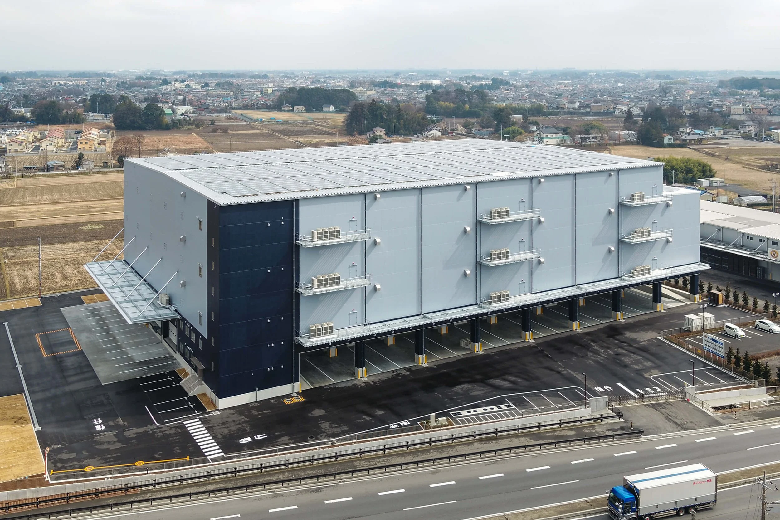 Large modern warehouse building with solar panels on the roof, multiple air conditioning units on the exterior walls, parking lot in front, located beside a highway, with a town and fields in the background.