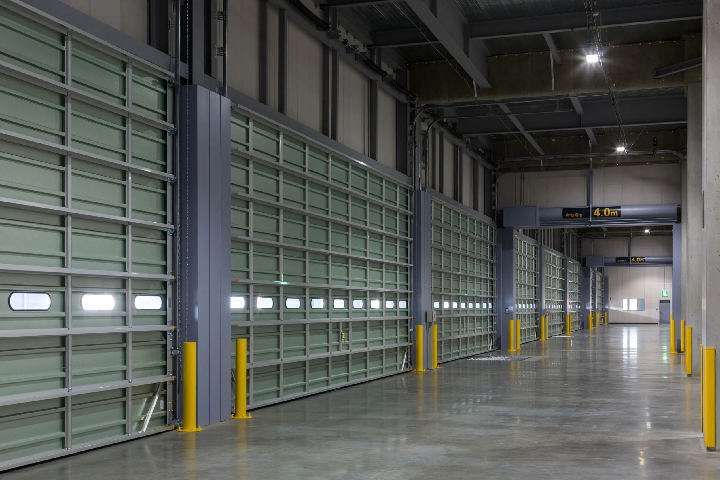 Empty industrial warehouse with large green rolling garage doors, concrete floor, yellow safety bollards, and overhead clear height limit signs of 4.0 meters.