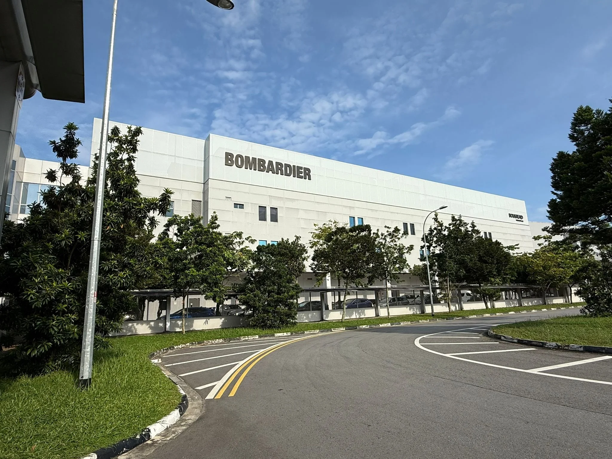Exterior of a large white building with the word 'Bombardier' on it, surrounded by trees, a parking lot, and a curved driveway under a partly cloudy blue sky.