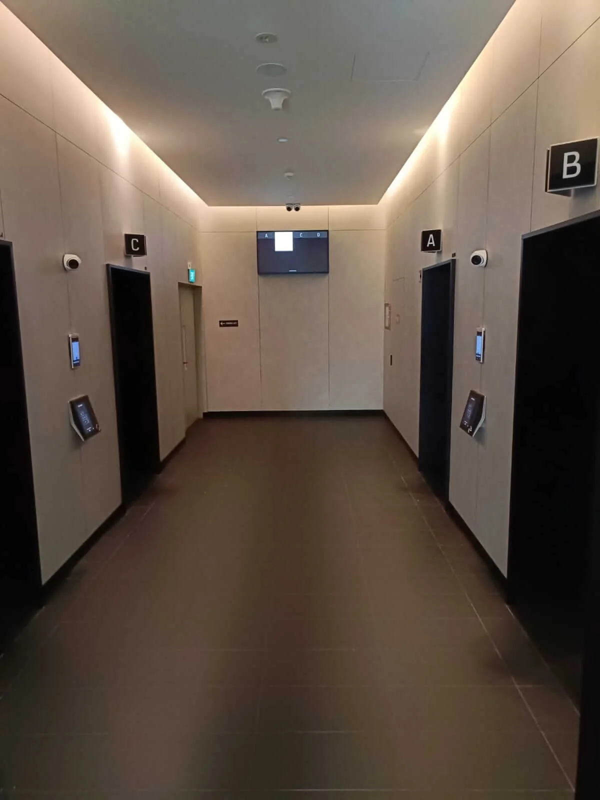 A hallway with four elevator doors labeled A, B, C, and D, each with a small touchscreen panel beside them, and a dark tiled floor.