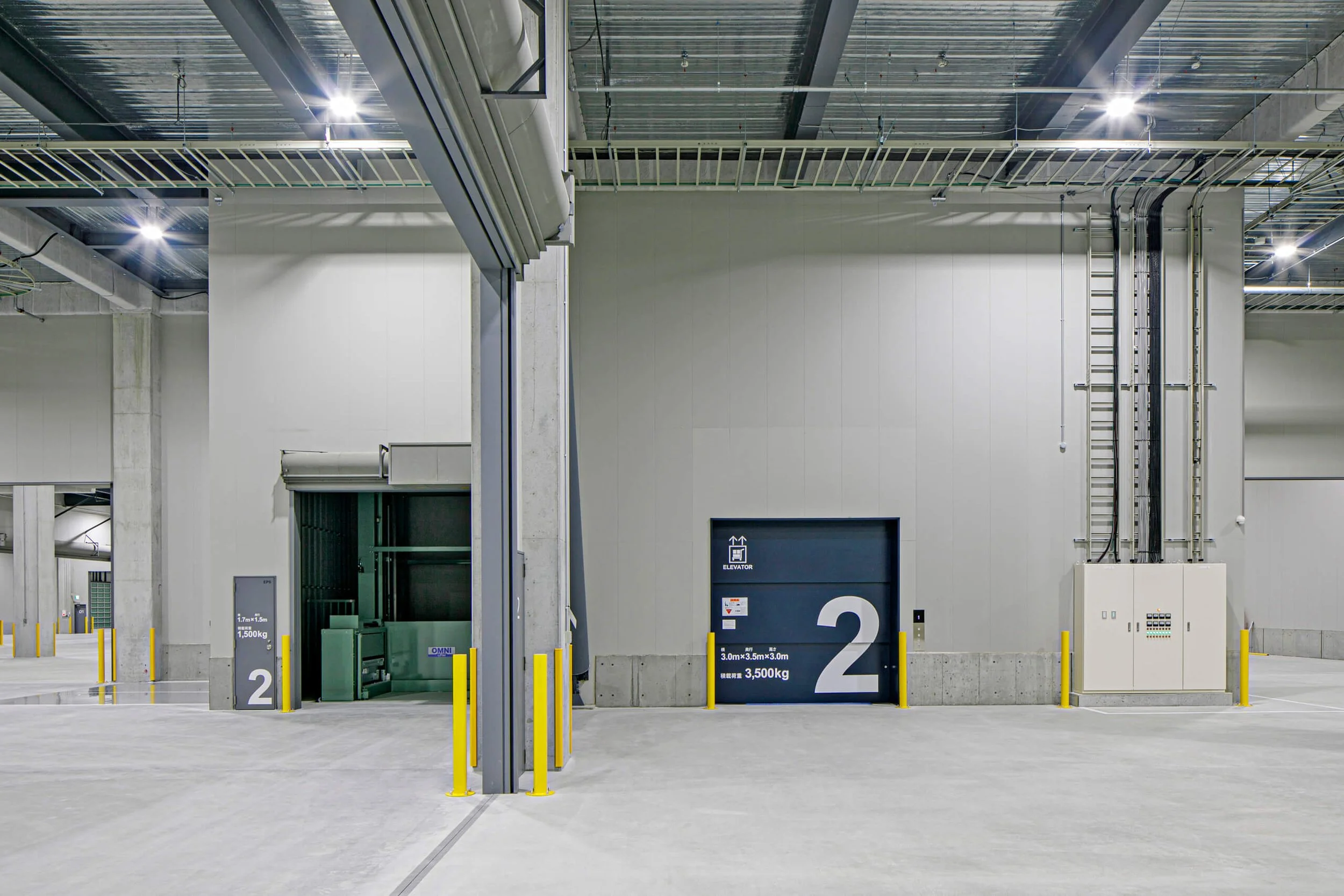 Empty industrial warehouse with a closed loading dock door marked with the number 2, overhead lighting, concrete walls and floors, and exposed ceiling piping.