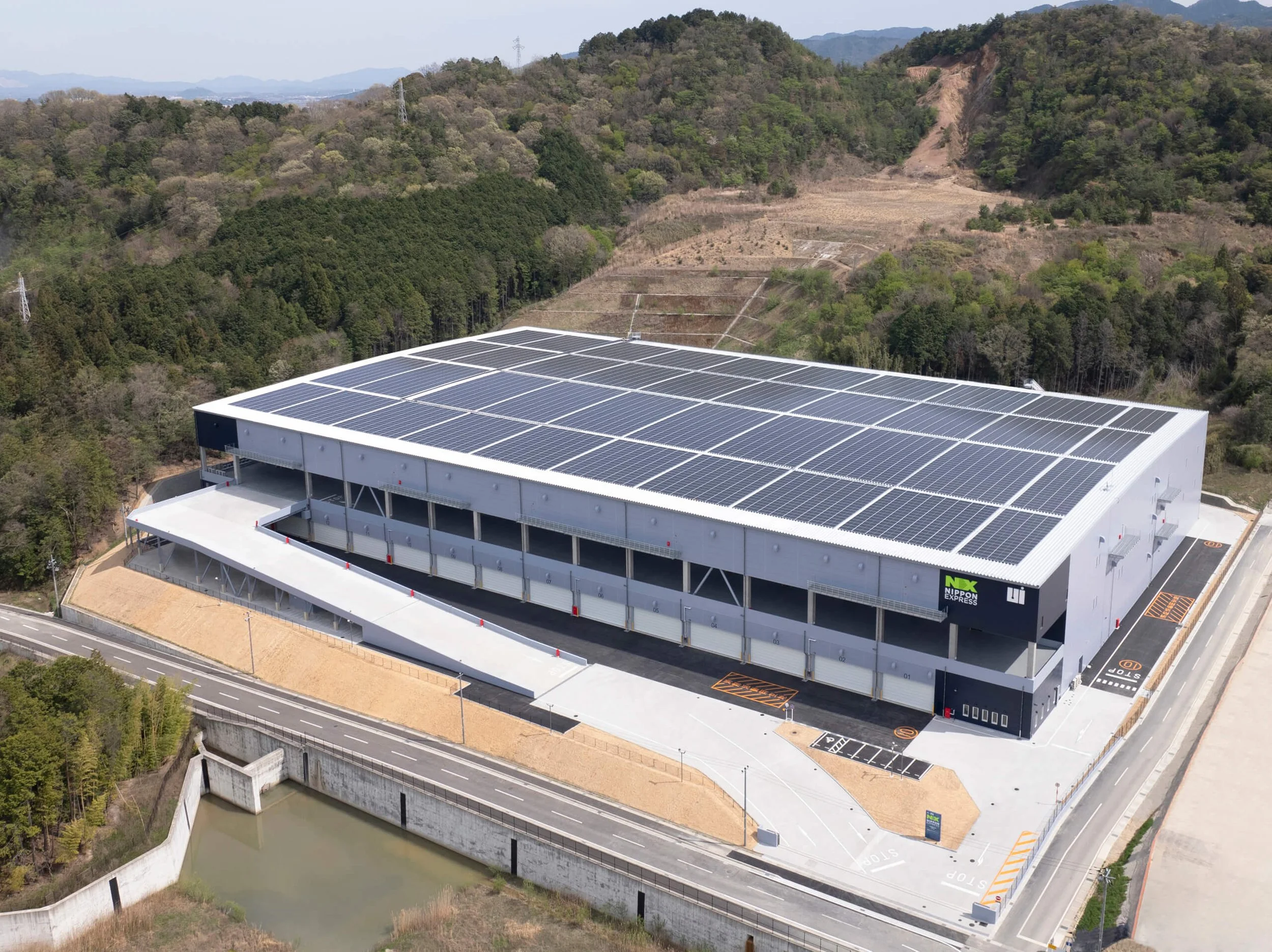 A large commercial building with a rooftop covered in solar panels, situated in a hilly, semi-rural area with surrounding forest and mountains in the background.