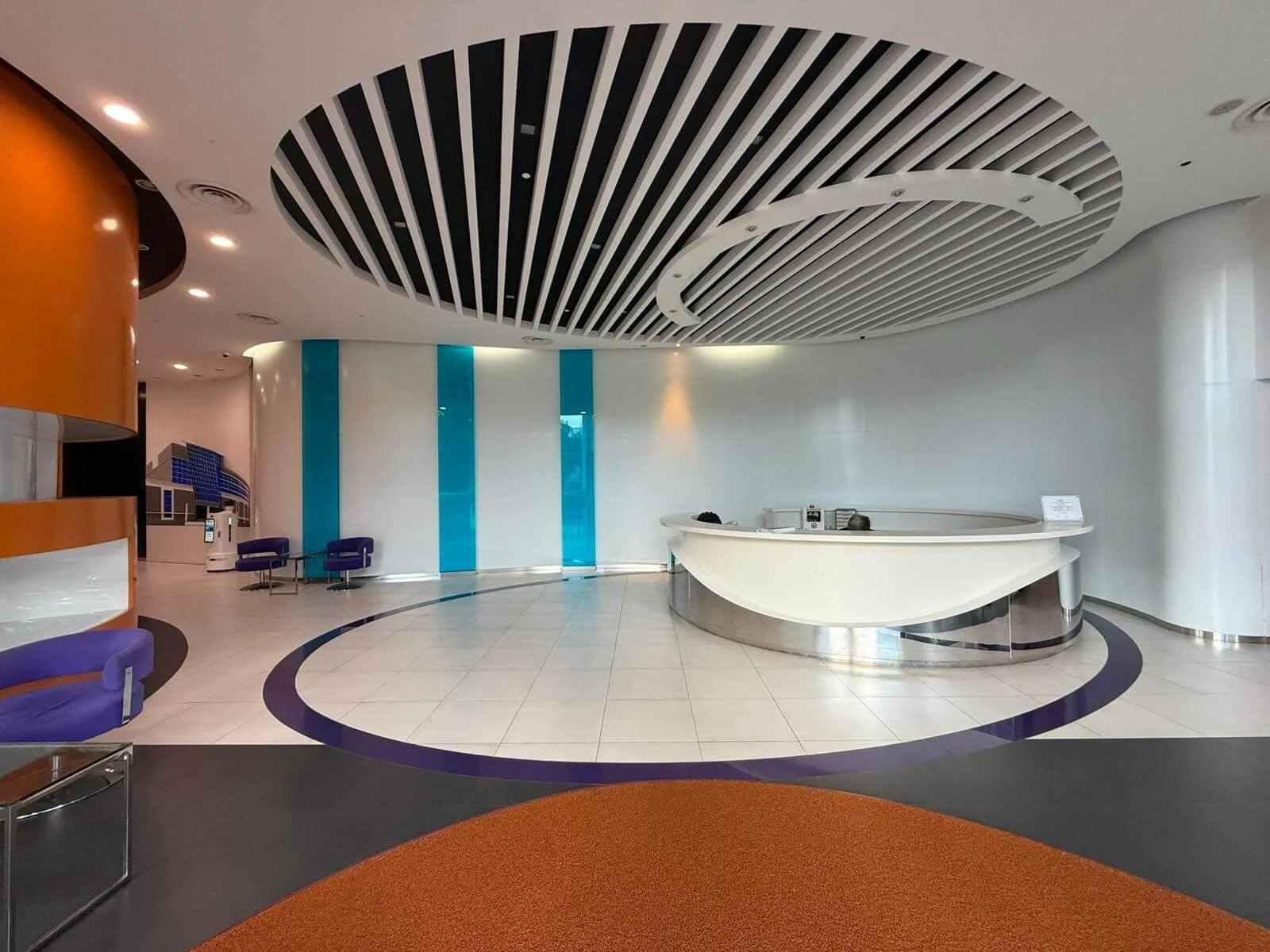 Modern hotel lobby with a curved white reception desk, purple chairs, orange carpet, and blue glass panels on the wall.