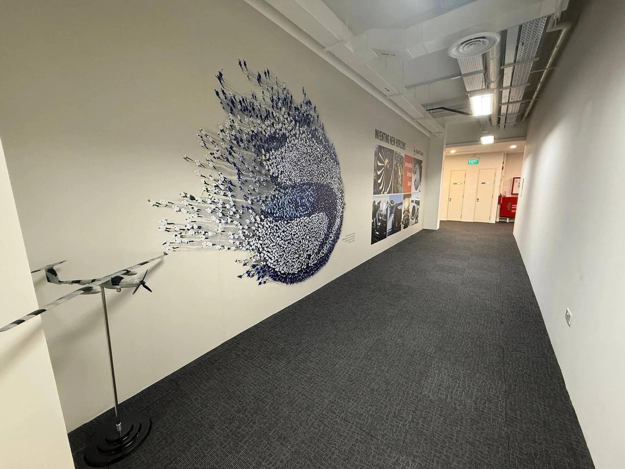 Hallway with wall display of an abstract swirling pattern of small, colored, human-shaped figures and informational posters about manufacturing innovations. Model airplane sculpture on stand on the left side.
