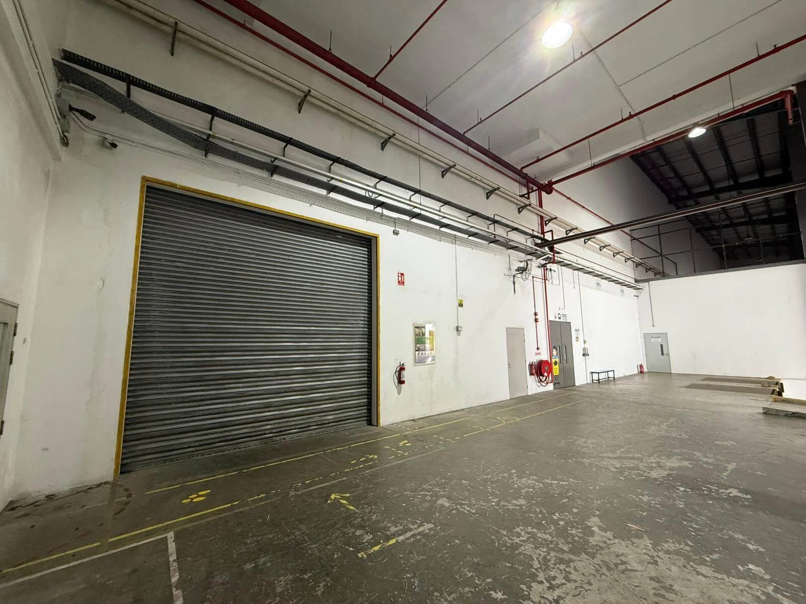 Empty industrial warehouse with a large metal rolling door, fire safety equipment, and concrete floor.