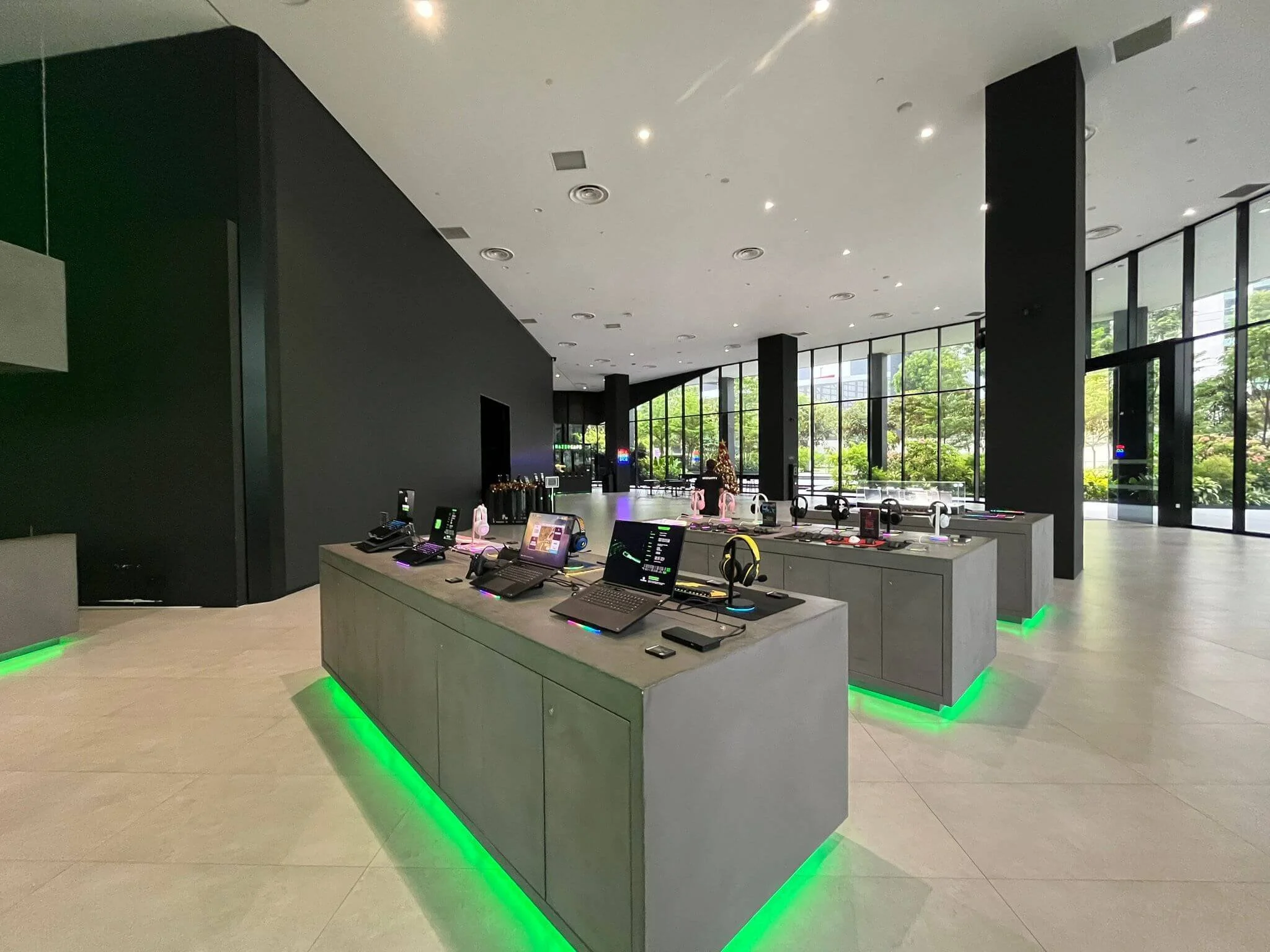 Modern electronics store interior with laptops, smartphones, and accessories on display, illuminated with green lighting accents at the base of gray display counters, large floor-to-ceiling windows allowing natural light, and black walls and pillars.
