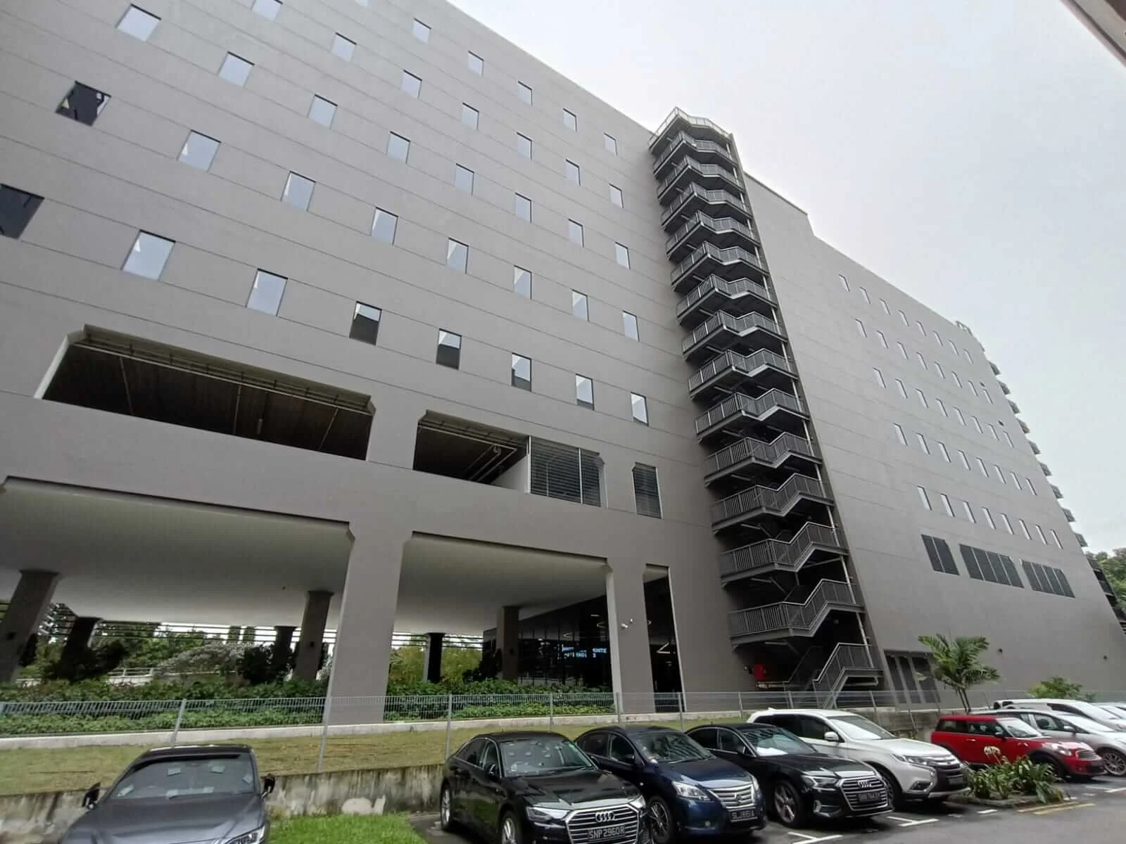 Tall modern building with multiple floors, external staircase, parking lot with cars, greenery in front.
