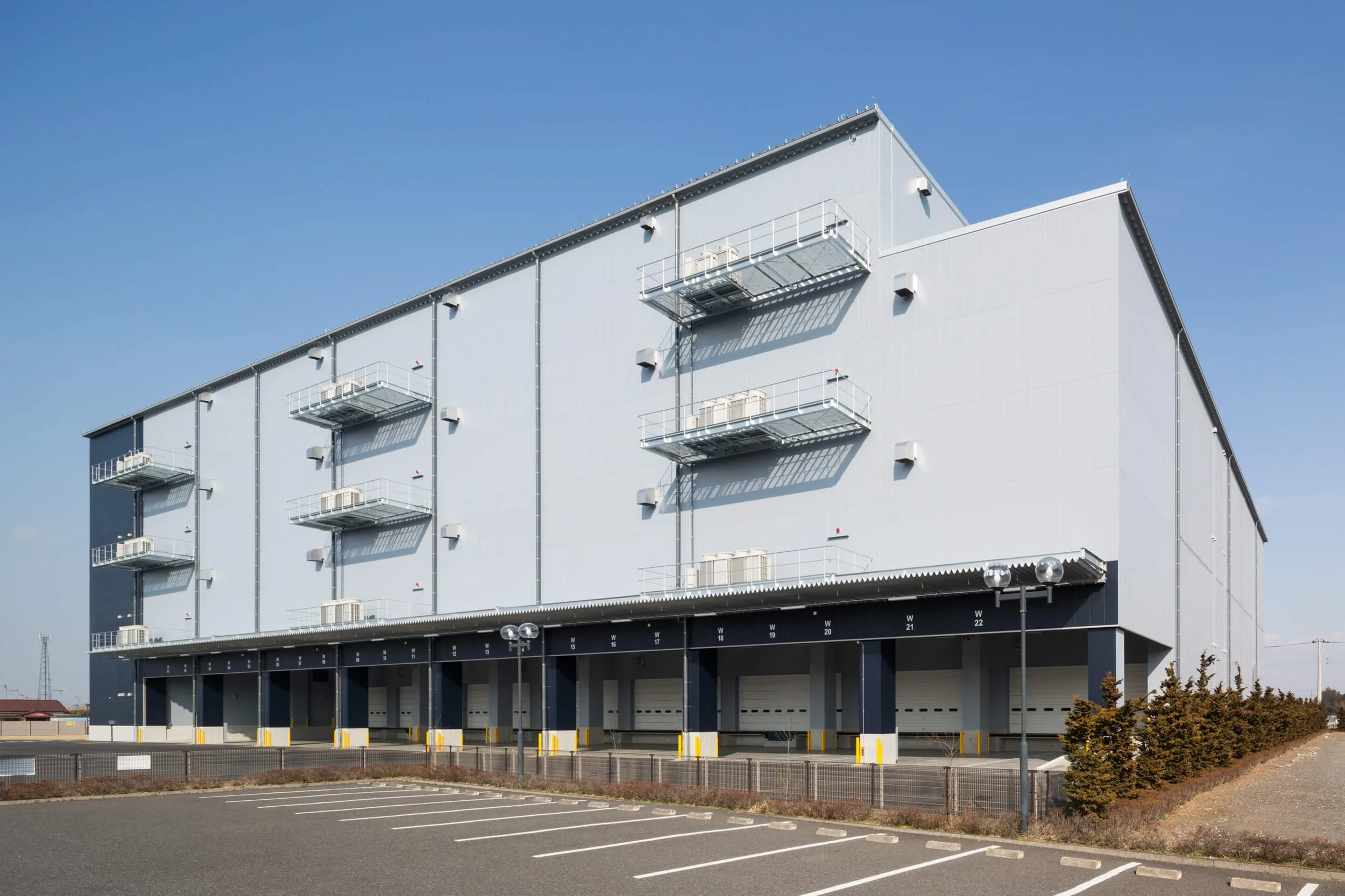 Large industrial warehouse building with multiple loading docks and a parking lot in the foreground under a clear blue sky.