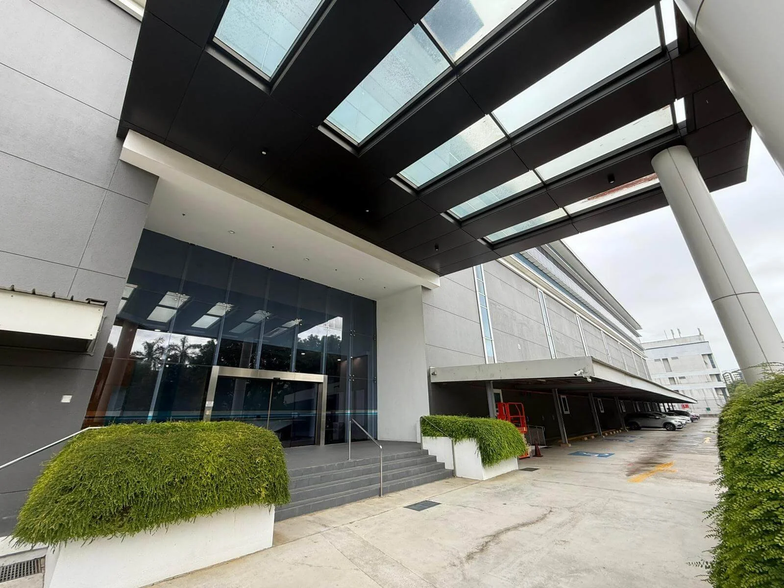 Modern multi-story building with a glass entrance, covered parking area with several cars, and landscaped bushes outside.