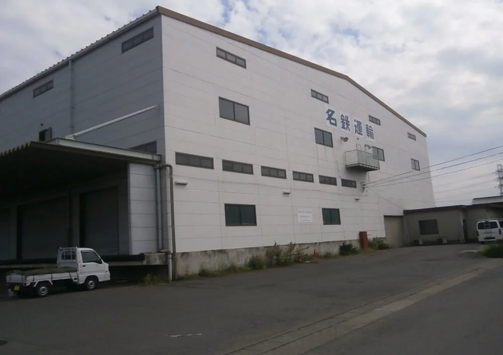 Large white industrial building with Japanese characters on the facade, small vans parked outside, and a cloudy sky overhead.