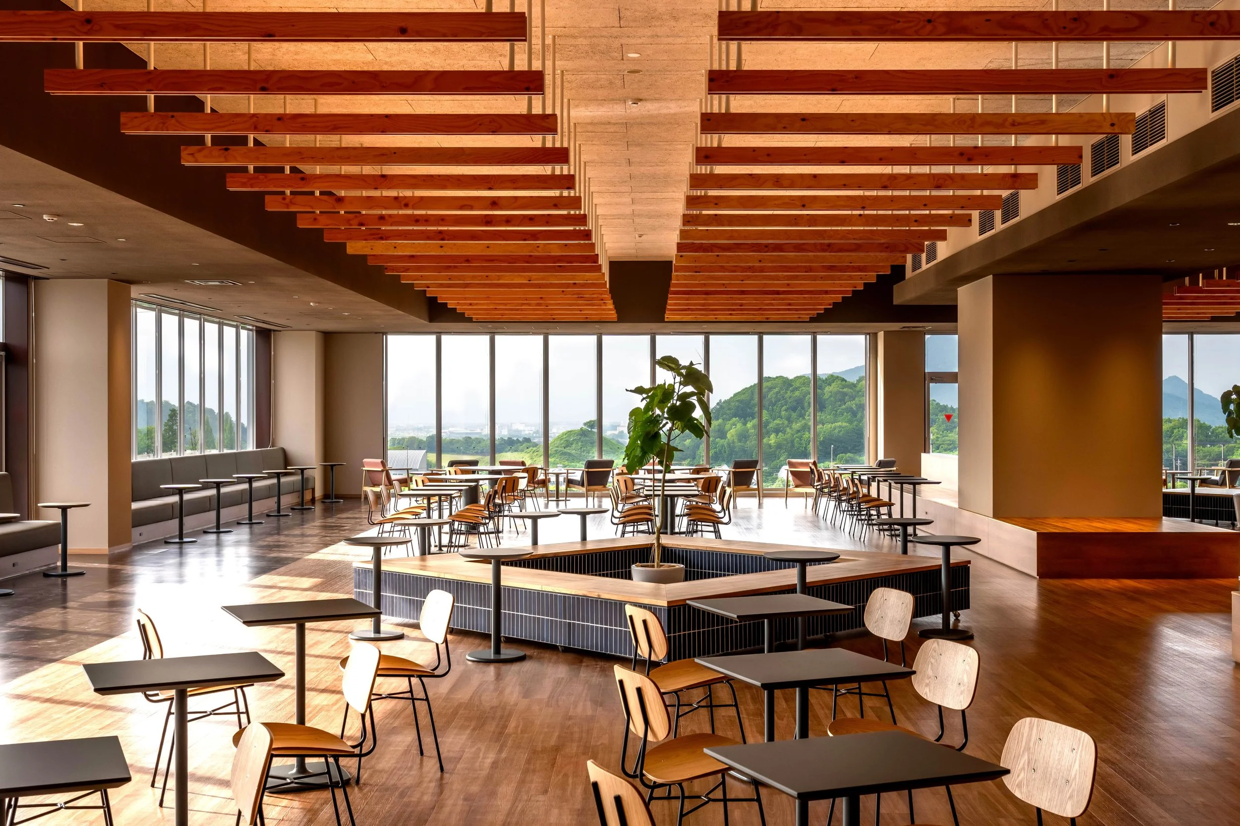 Modern, spacious dining area with wooden ceilings, large windows showing green hills, and a central planter with a tree surrounded by tables and chairs.