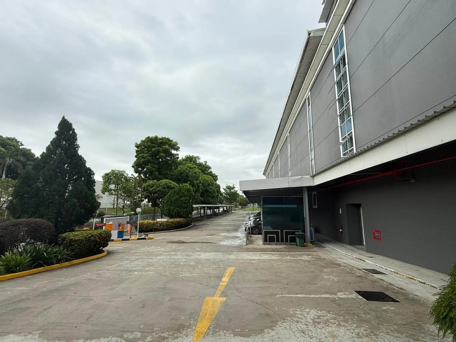 Empty parking lot next to a gray building with trees and cloudy sky in background.