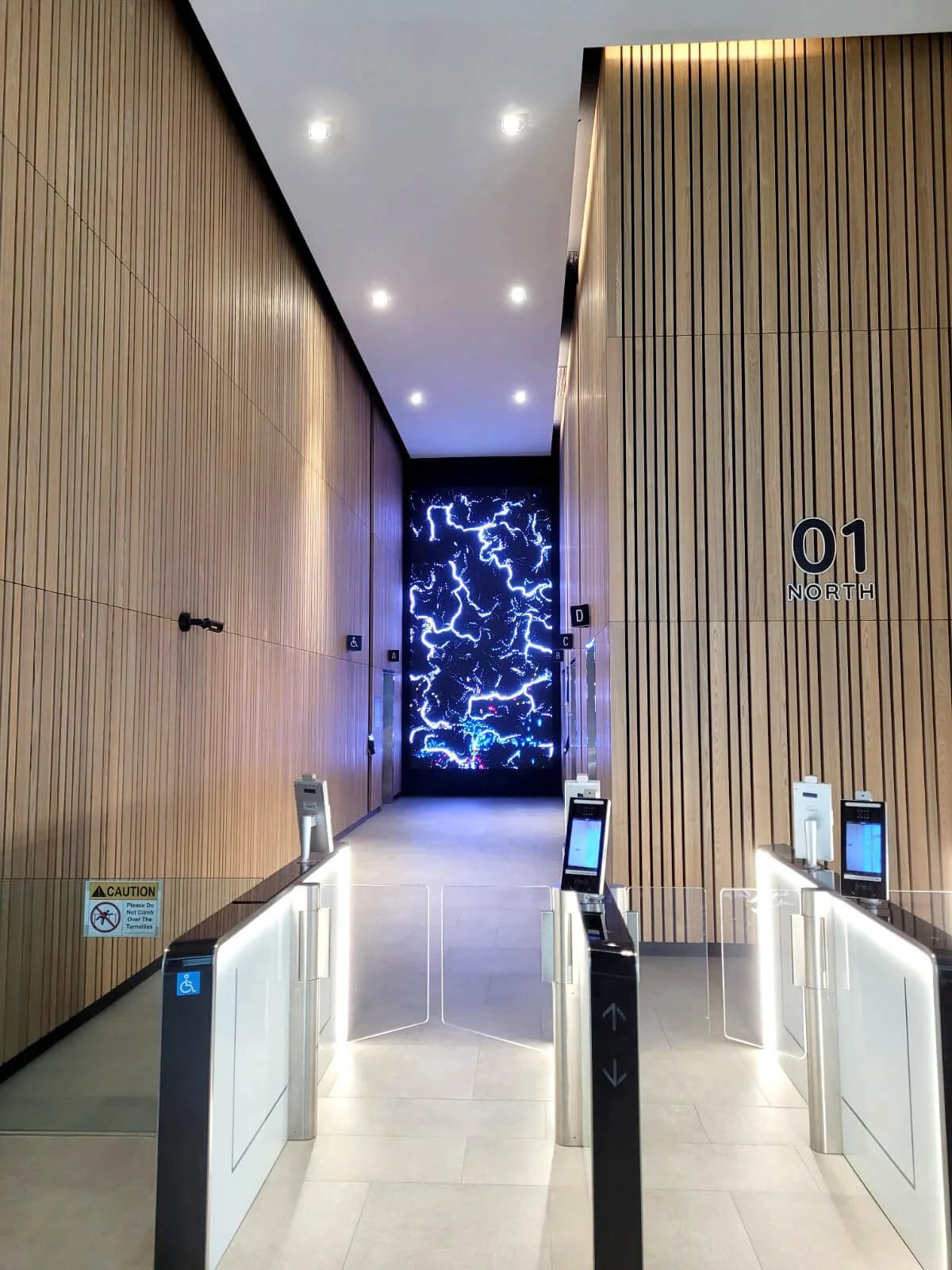 Indoor elevator lobby with wooden wall panels, ceiling lights, and a digital display at the far end with a lightning pattern. There are automatic gate barriers at the entrance with digital screens, and signage indicating the elevator is going north f