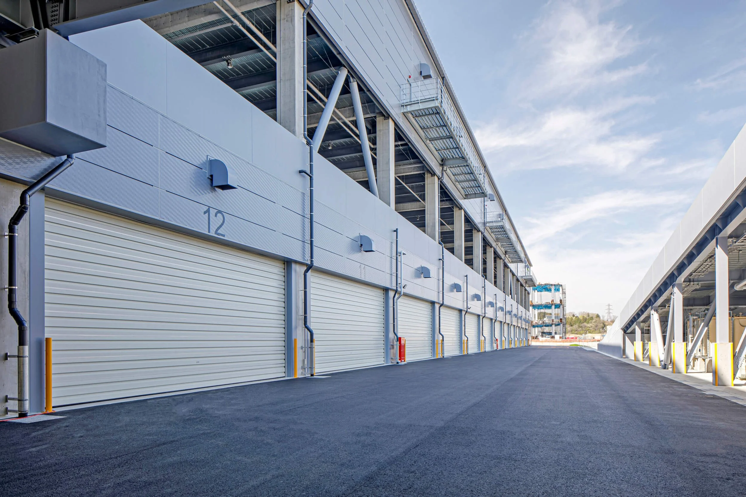 Exterior view of a modern parking garage with multiple garage bays, marked with number 12, and an empty asphalt driveway, under a partly cloudy sky.