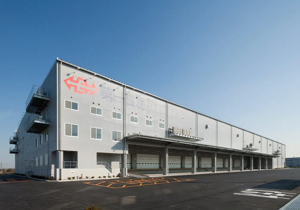 Large white warehouse with multiple loading docks, small windows, a fire escape balcony on the left, and red and gray graphic on the top left corner, under a clear blue sky.