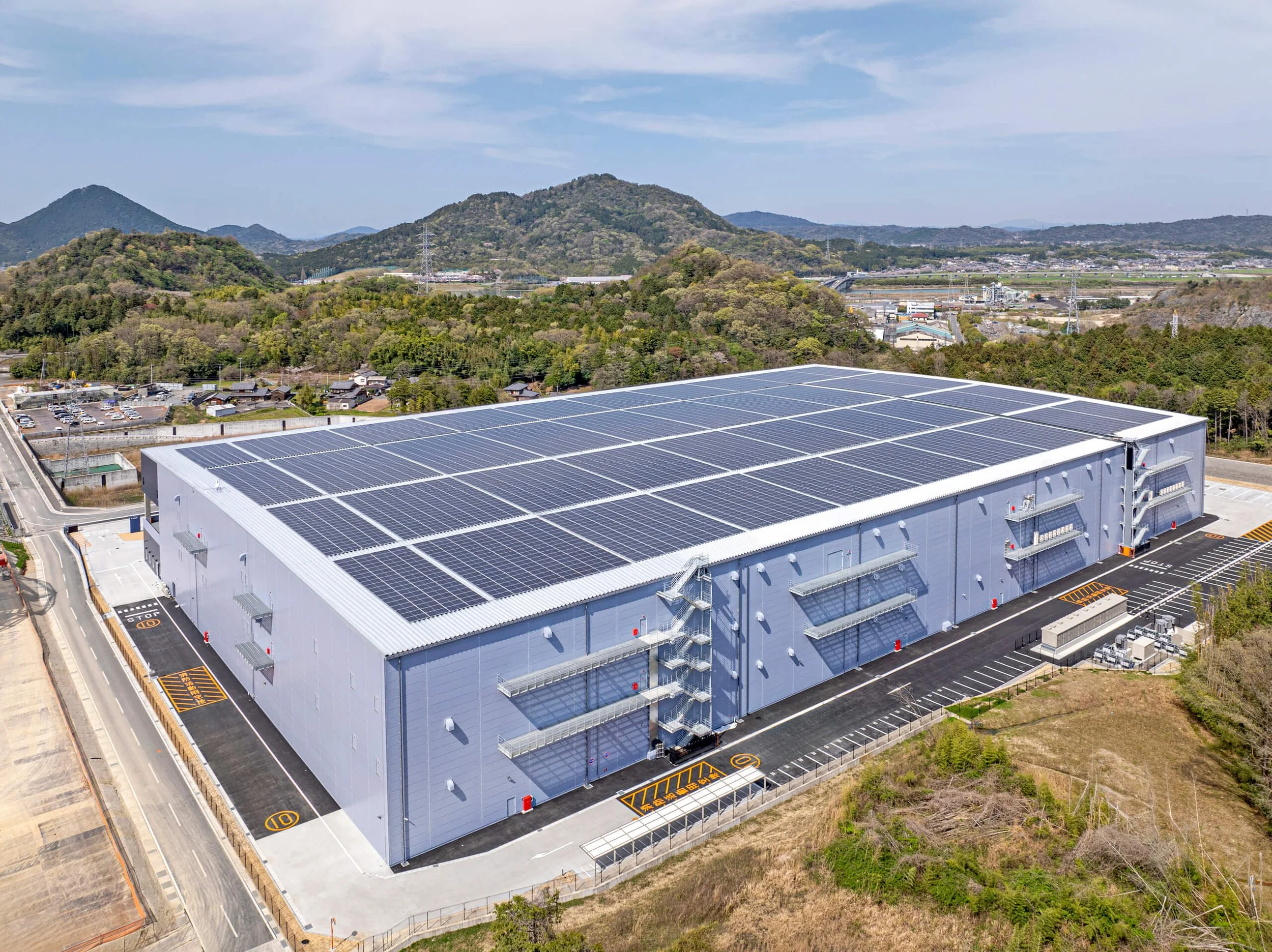 Large industrial building with a flat roof covered in solar panels, surrounded by parking lots and set in a mountainous area.