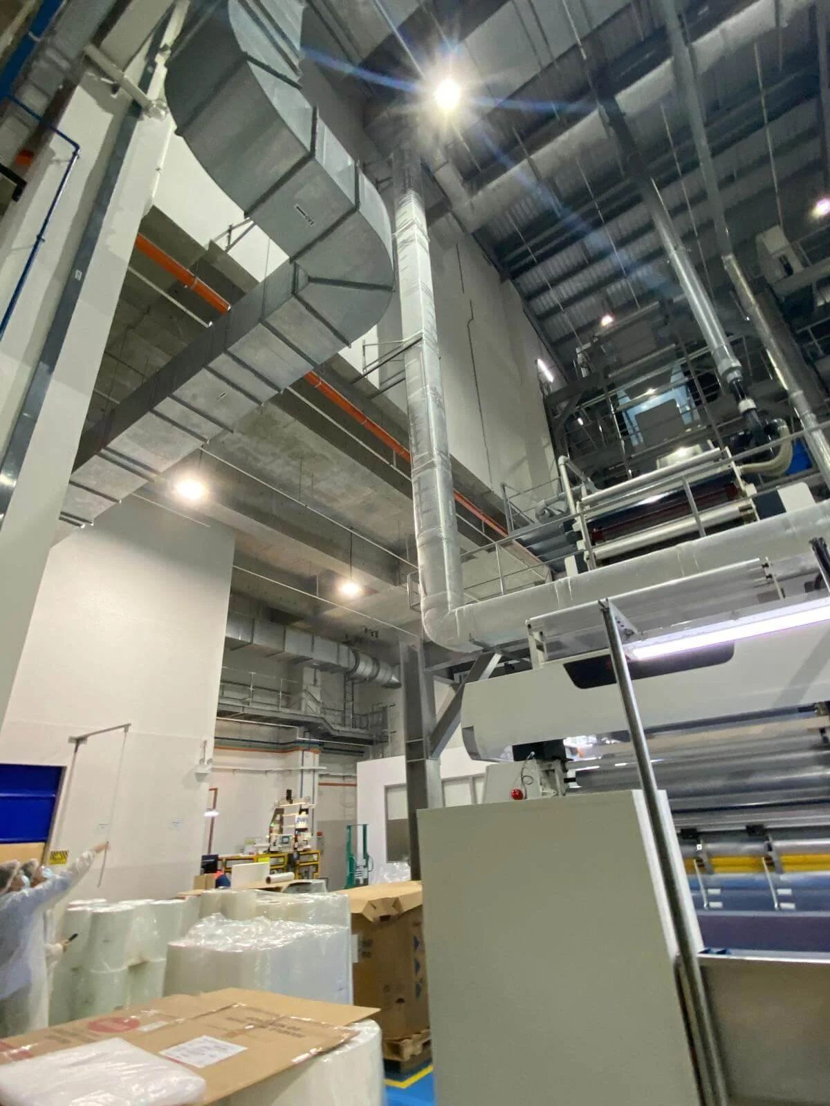 Inside an industrial factory with large ventilation ducts, machinery, and stacks of packaged products.