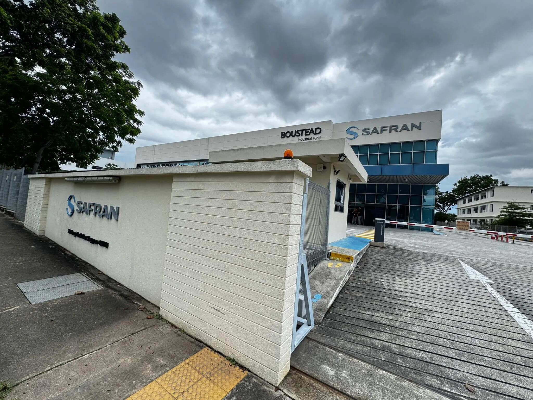 Exterior view of a modern office building with signs displaying 'SAFRAN,' 'BOUSTEAD,' and 'Industrial Fund' under a cloudy sky.