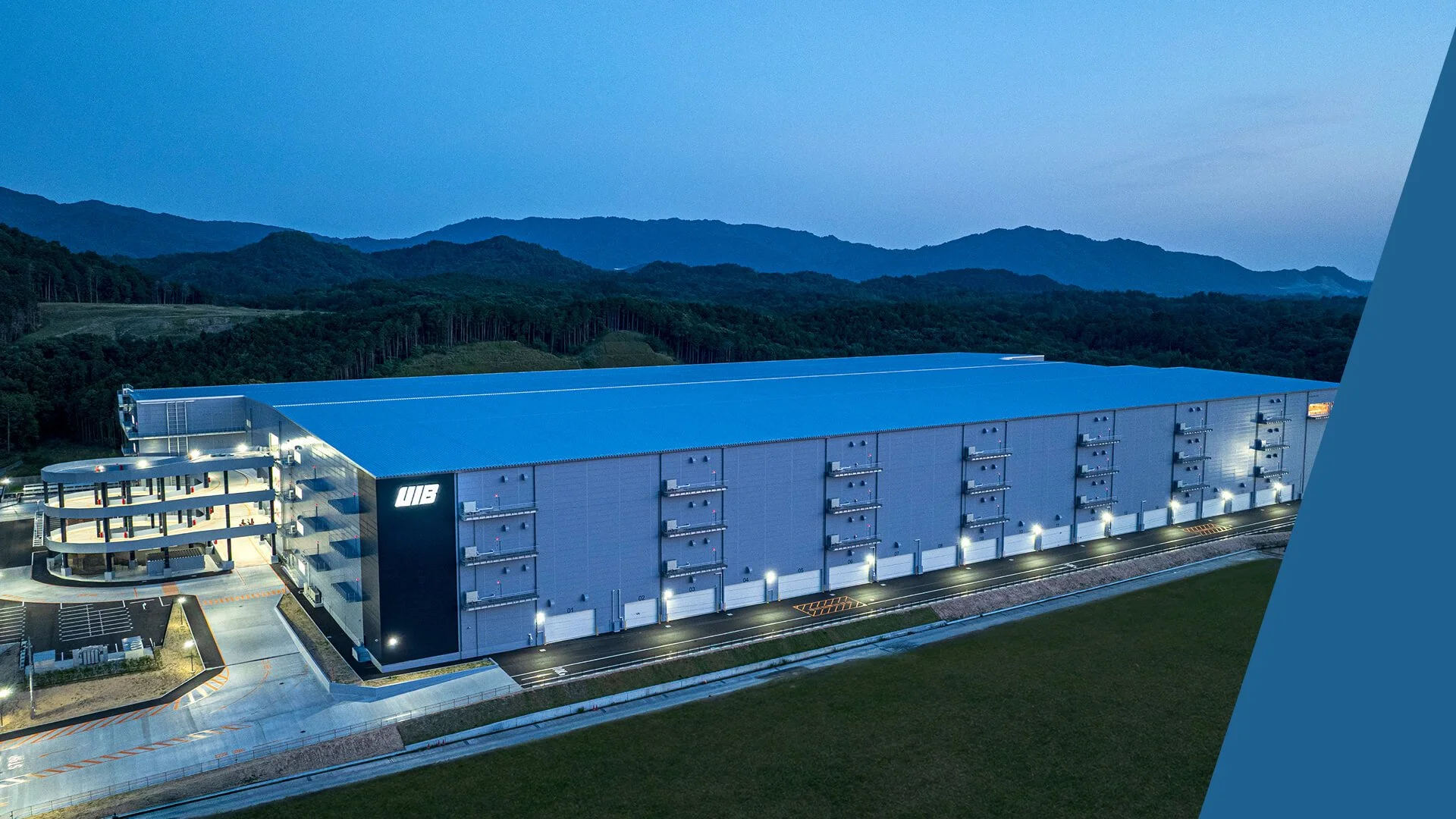 Large industrial warehouse building with multiple loading docks, surrounded by mountains at dusk, with exterior lights on.